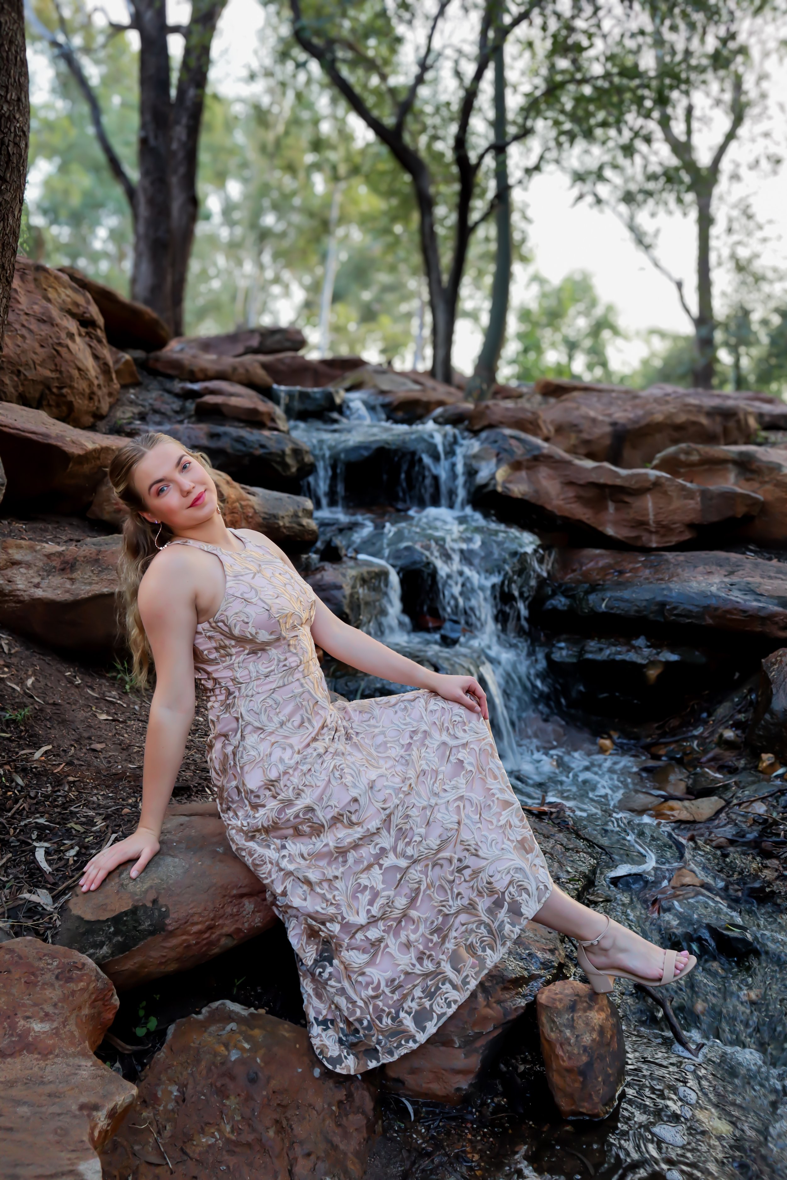 A woman in a long, light-colored dress sits on rocks beside a small waterfall in a wooded area.