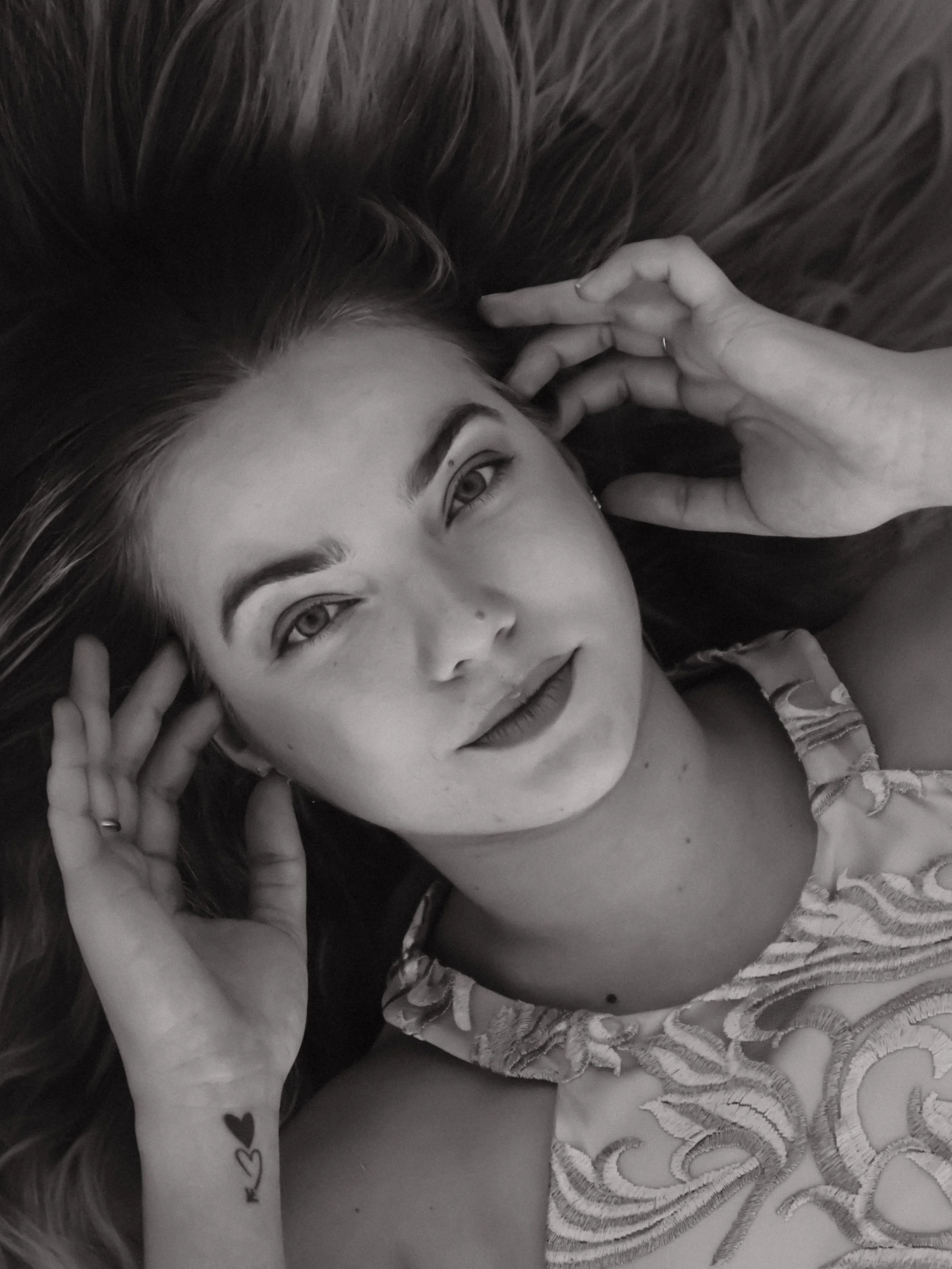 A young woman with long hair lying on a surface, looking into the camera, with her hands near her face, one hand resting on her temple, wearing a sleeveless top with embroidered patterns, and a small tattoo of hearts on her wrist.