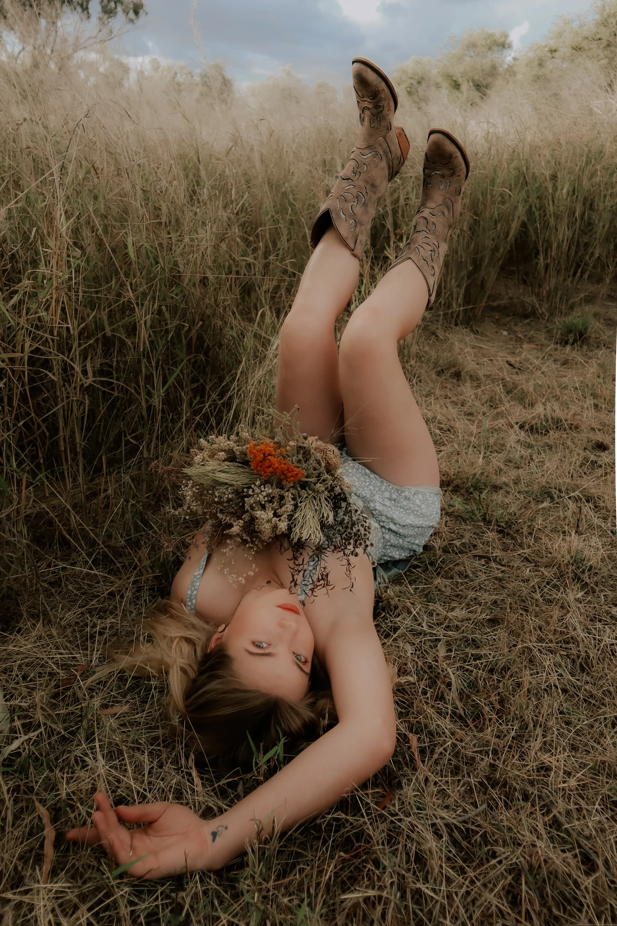 A woman lying in a field of dry grass, holding a bouquet of flowers, with her legs raised and wearing cowboy boots.