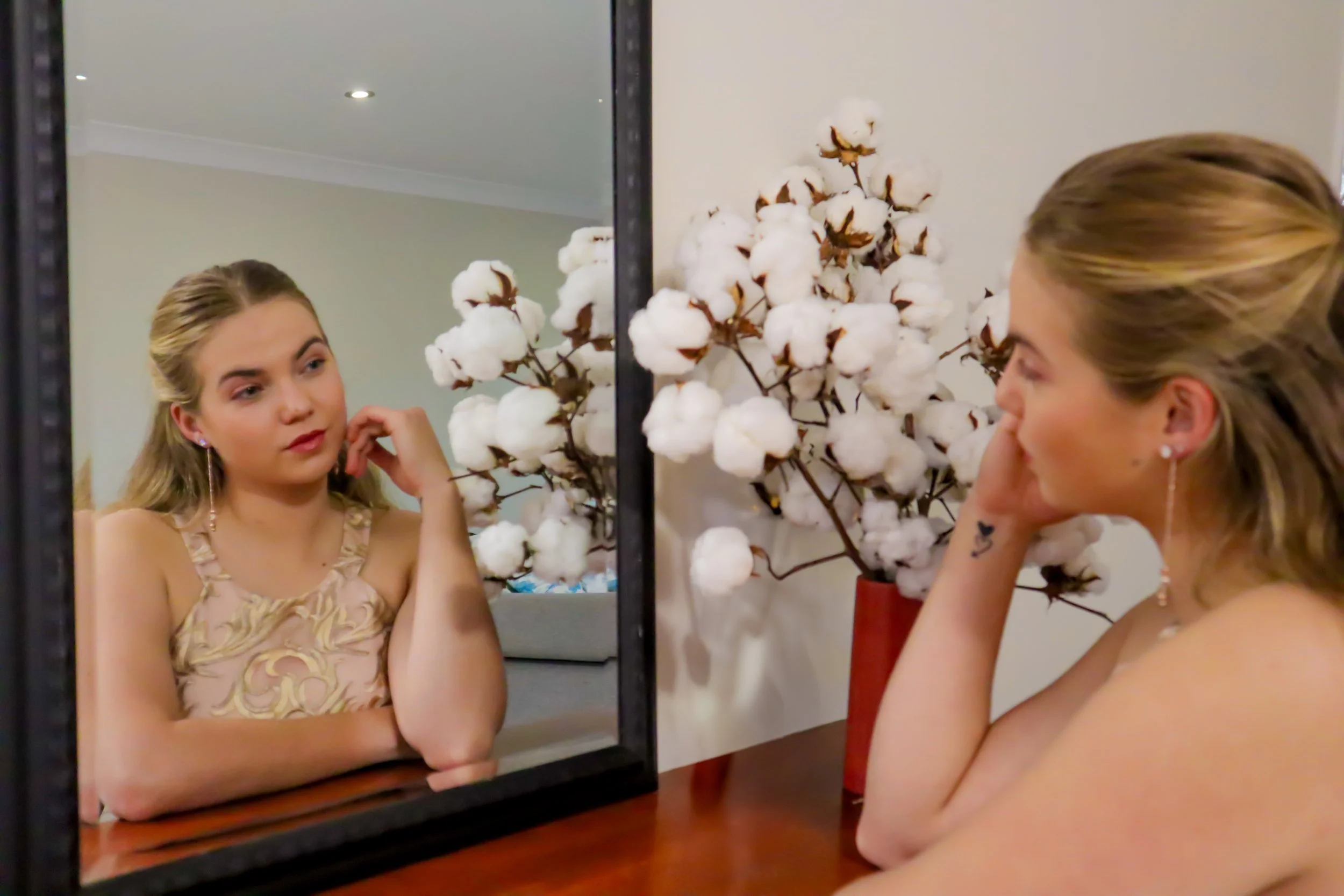 A woman looking at her reflection in the mirror with a cotton branch decoration and a red vase in the background.