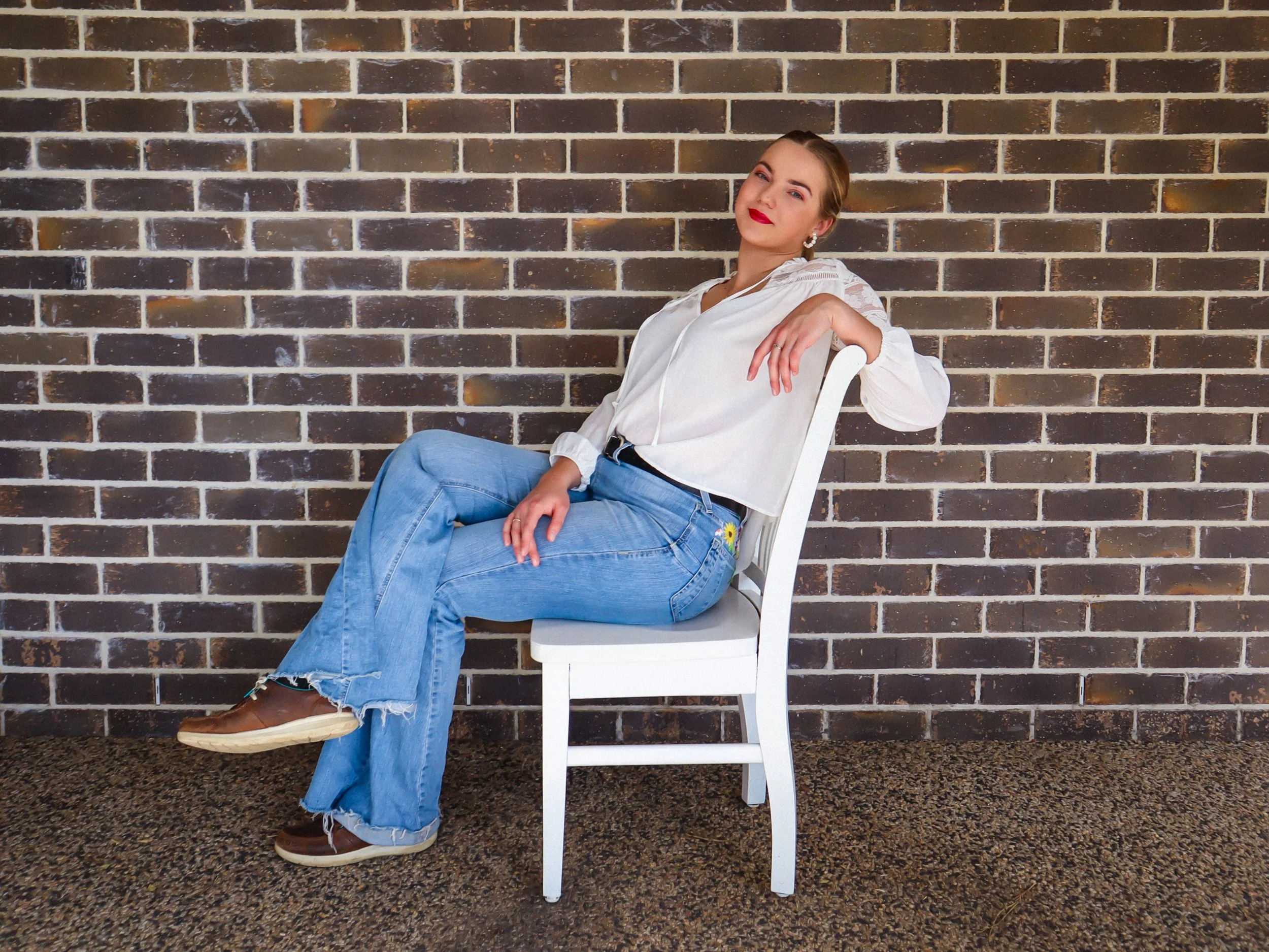 Woman in white blouse and ripped jeans sitting on a white chair against a brick wall, posing confidently with one arm resting on the chair and looking at the camera.