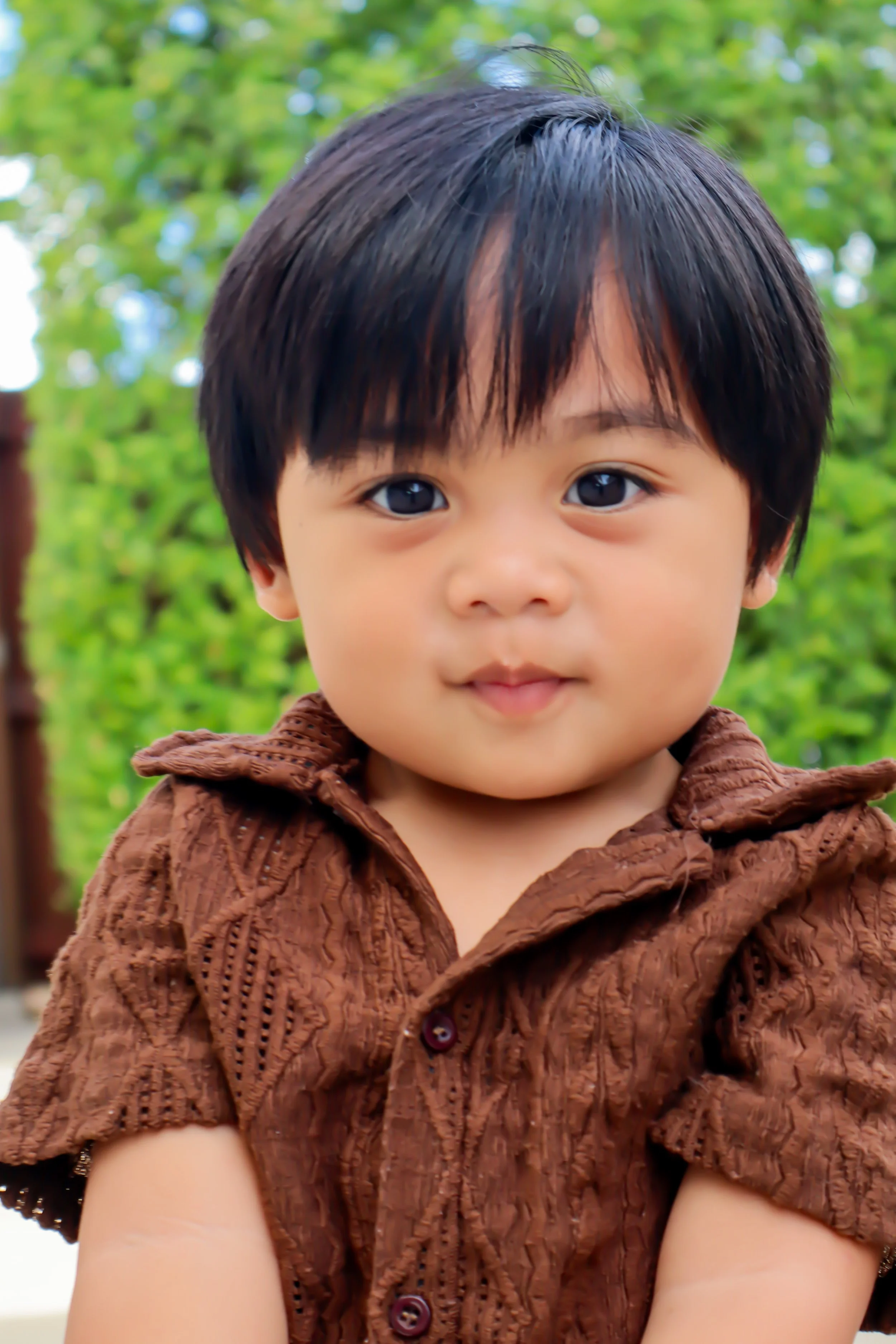 Close-up of a young child with dark hair, wearing a brown shirt, outdoors with green trees in the background.