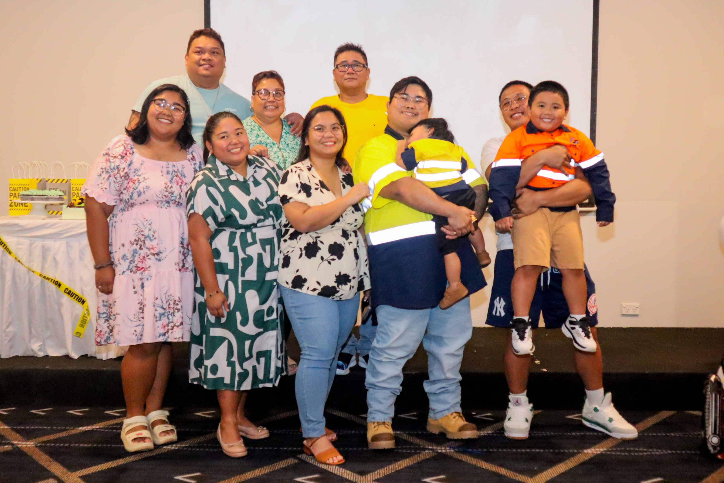 Group of people standing on stage, smiling, including children and adults, with some holding young children, in front of a white screen and table with caution tape.
