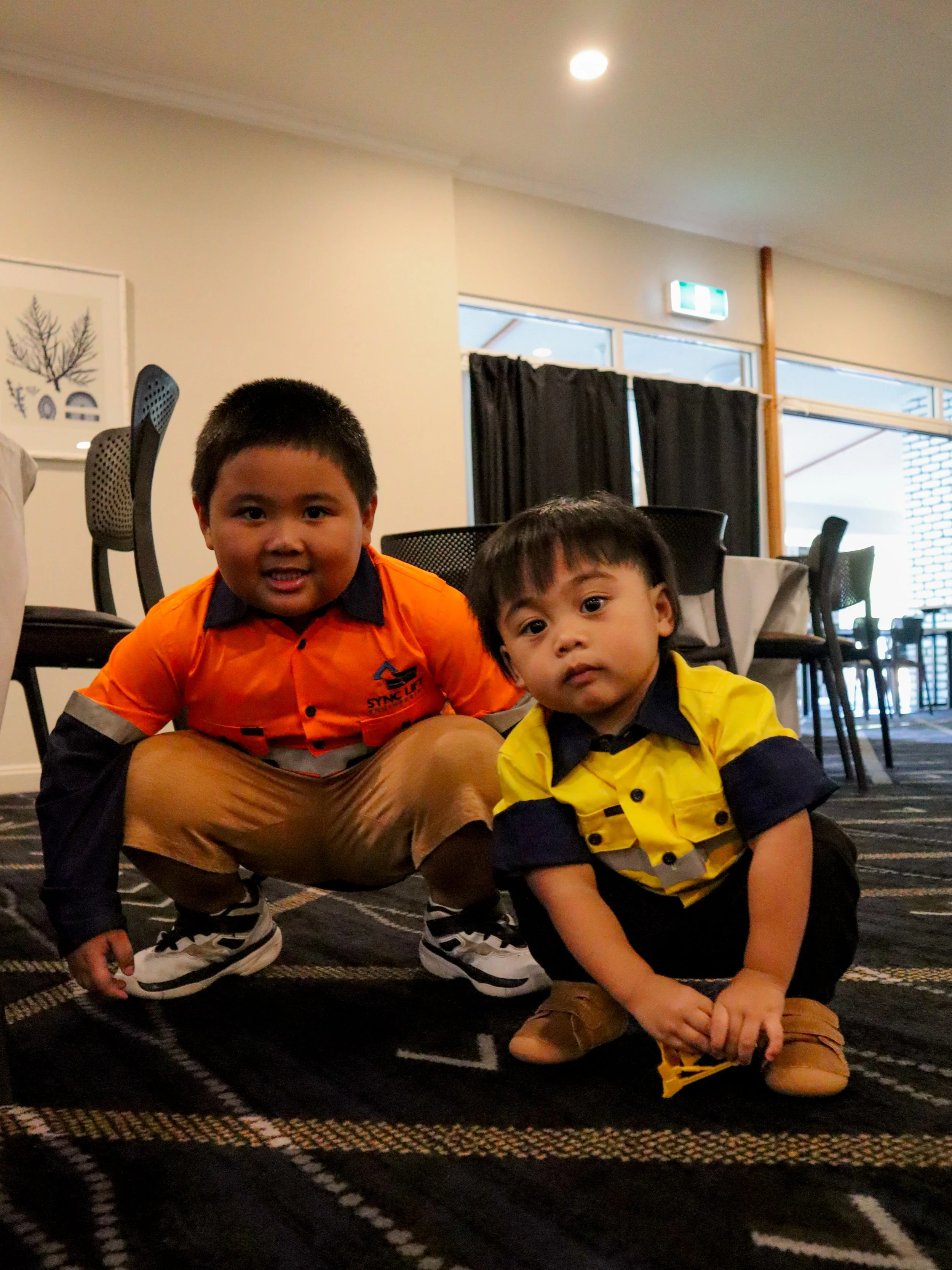 Two young boys crouch on a dark patterned carpet in a room with tables, chairs, and windows. Both boys wear colorful shirts, one in orange and the other in yellow, with dark collars and shoulder accents.