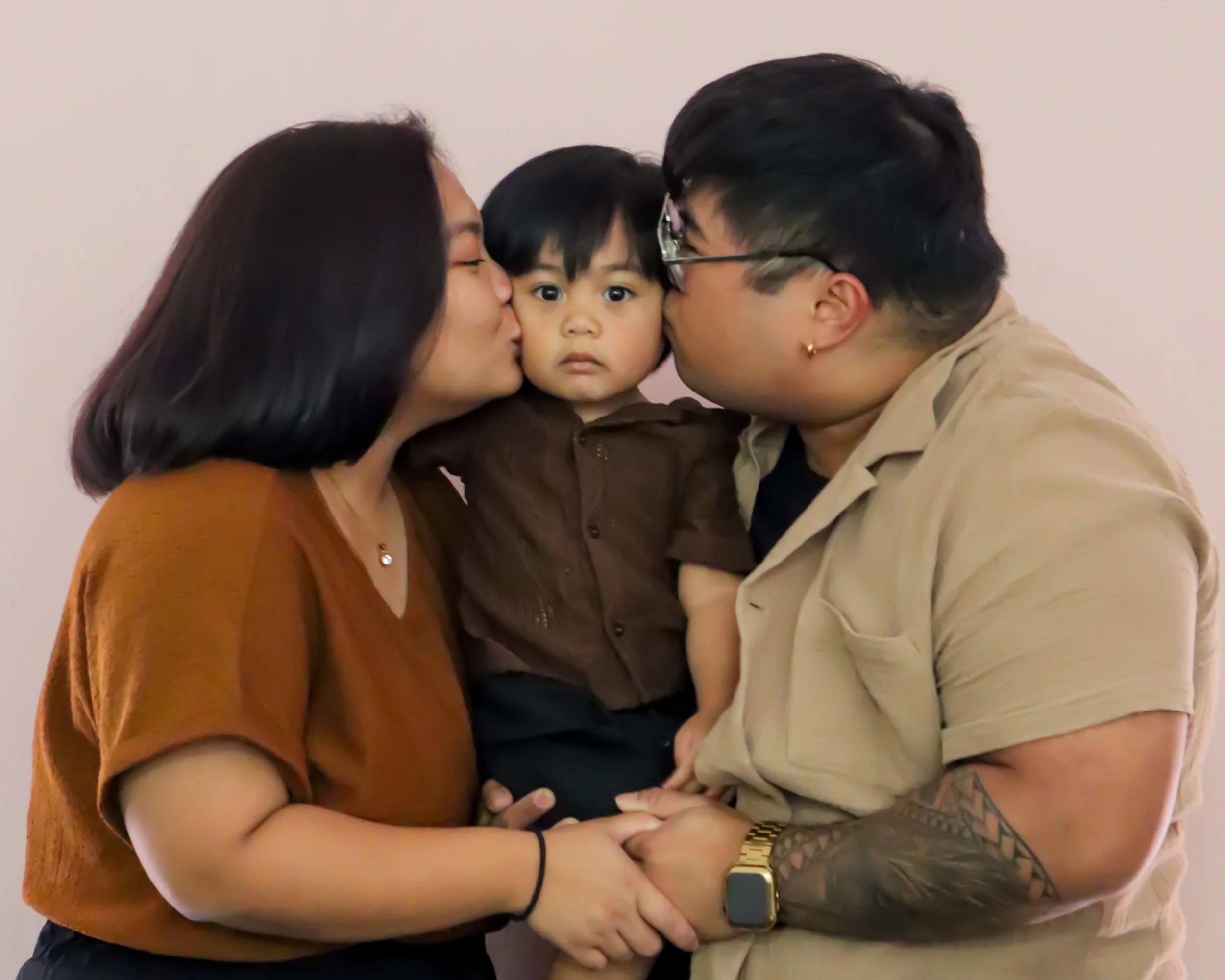 A young child being kissed on both cheeks by two adults, one woman and one man, in front of a plain light-colored wall.