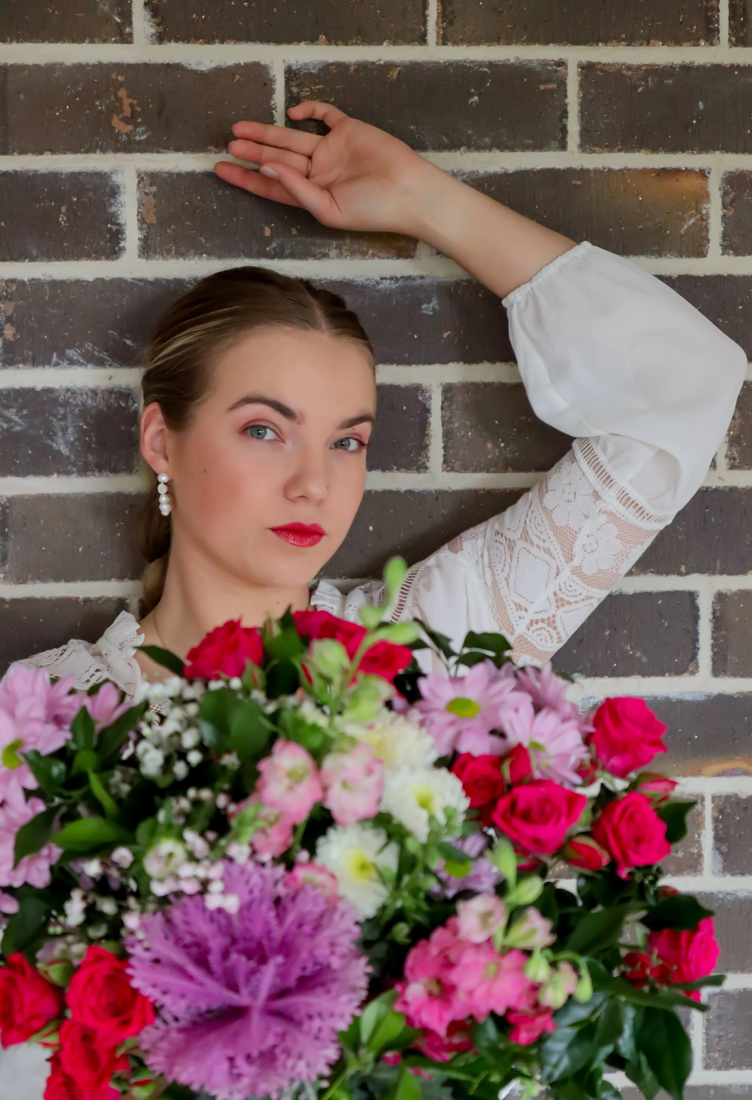 A woman with blue eyes and red lipstick, wearing pearl earrings and a white lace top, poses against a brick wall while holding a large bouquet of colorful pink, red, and white flowers.