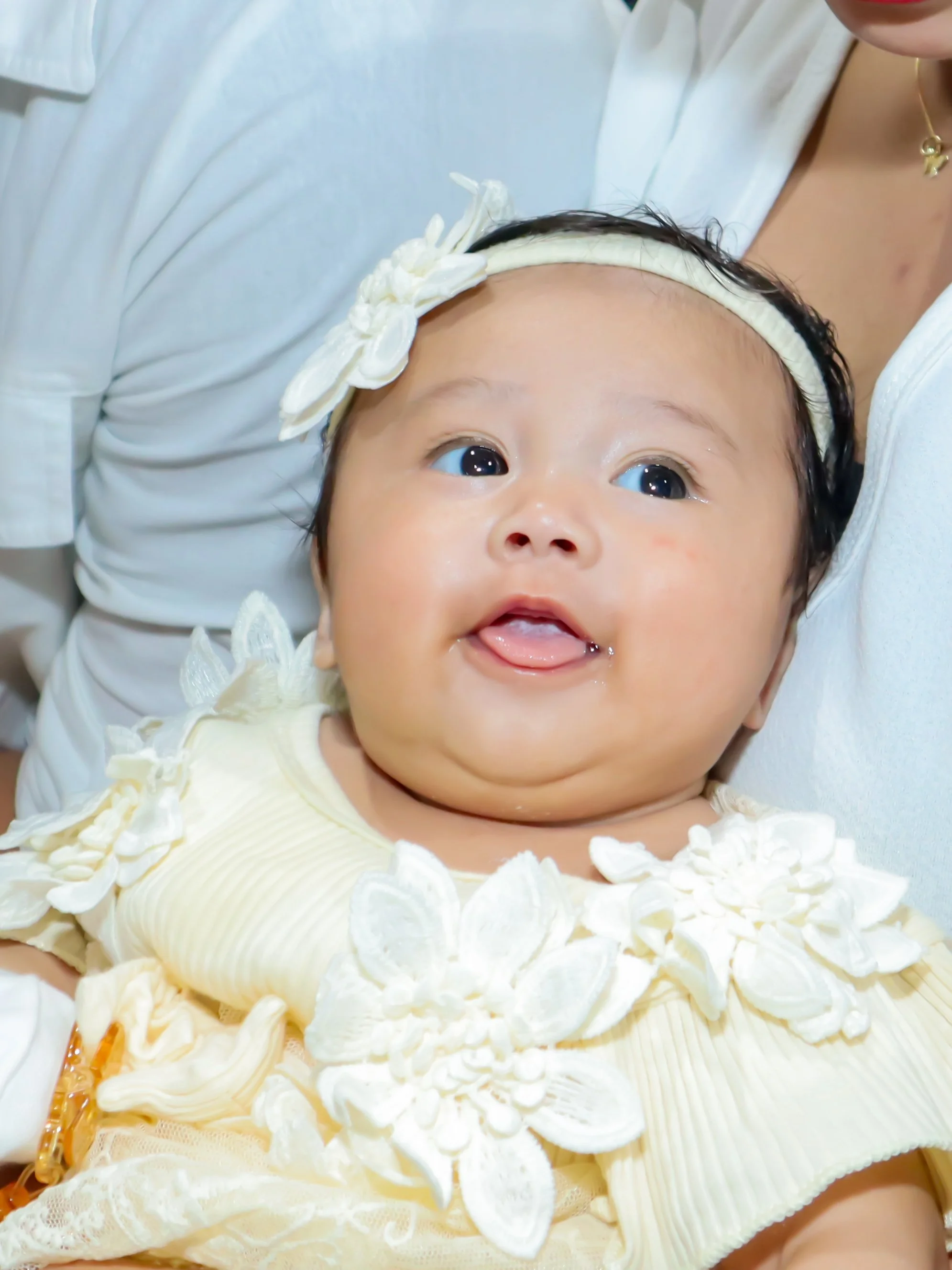 Close-up of a smiling baby girl wearing a cream-colored dress with floral embellishments and a headband, held by an adult.