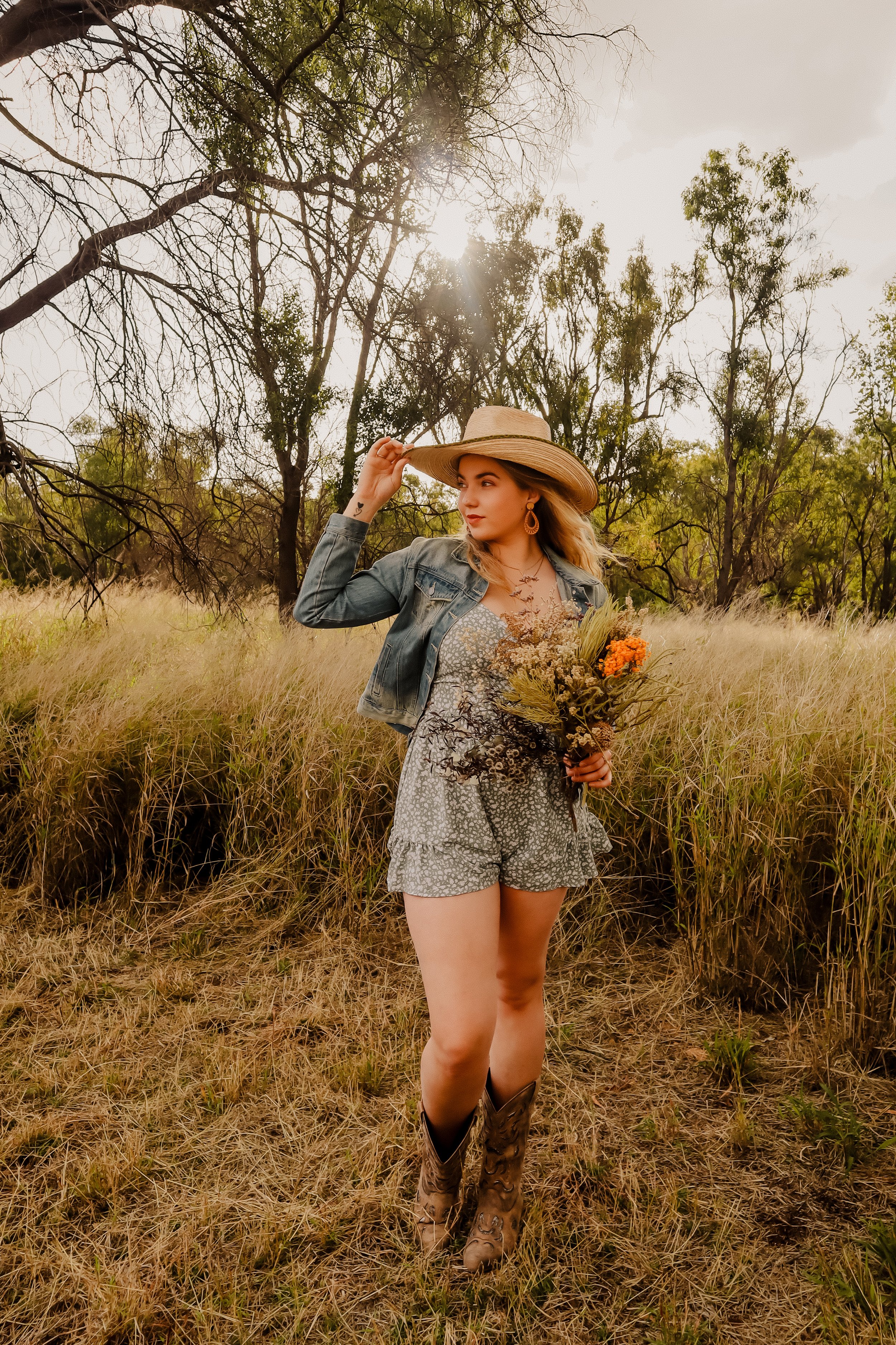 Young woman with blonde hair wearing a straw hat, denim jacket, floral romper, and cowboy boots, holding a bouquet of wildflowers in a field with tall grass and trees, sunlight filtering through the branches.