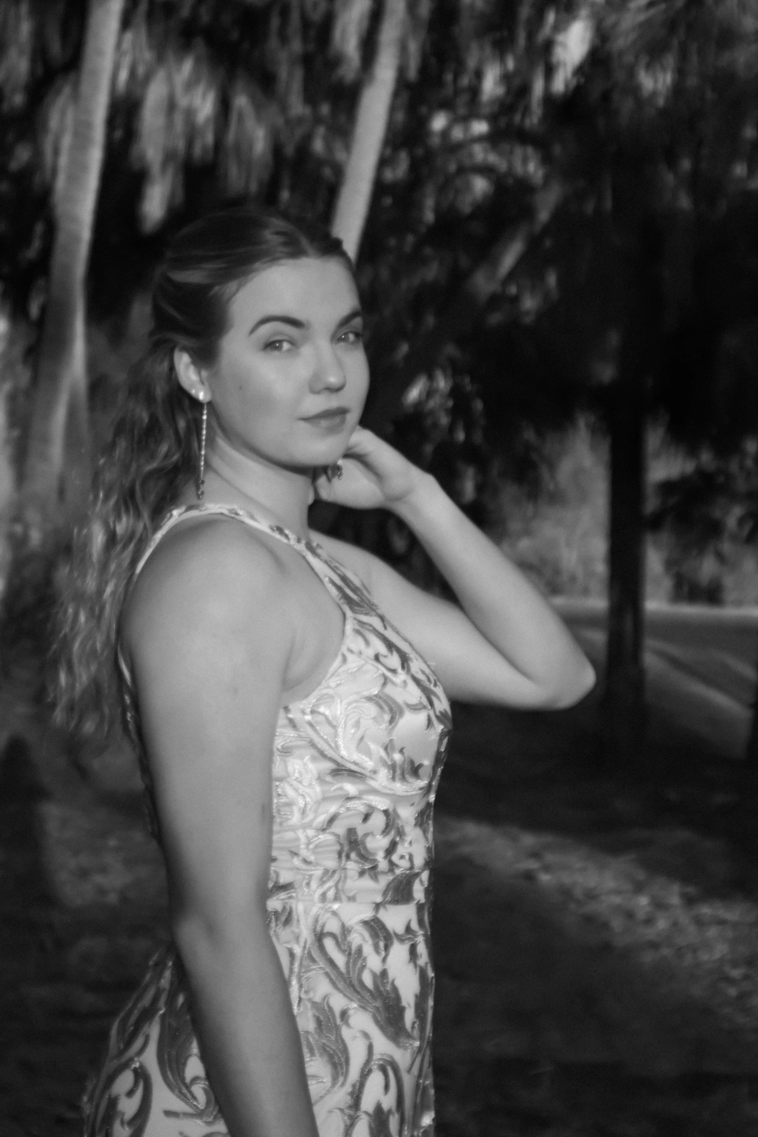 A young woman with wavy hair wearing a patterned sleeveless dress, standing outdoors at night with trees and a path in the background.