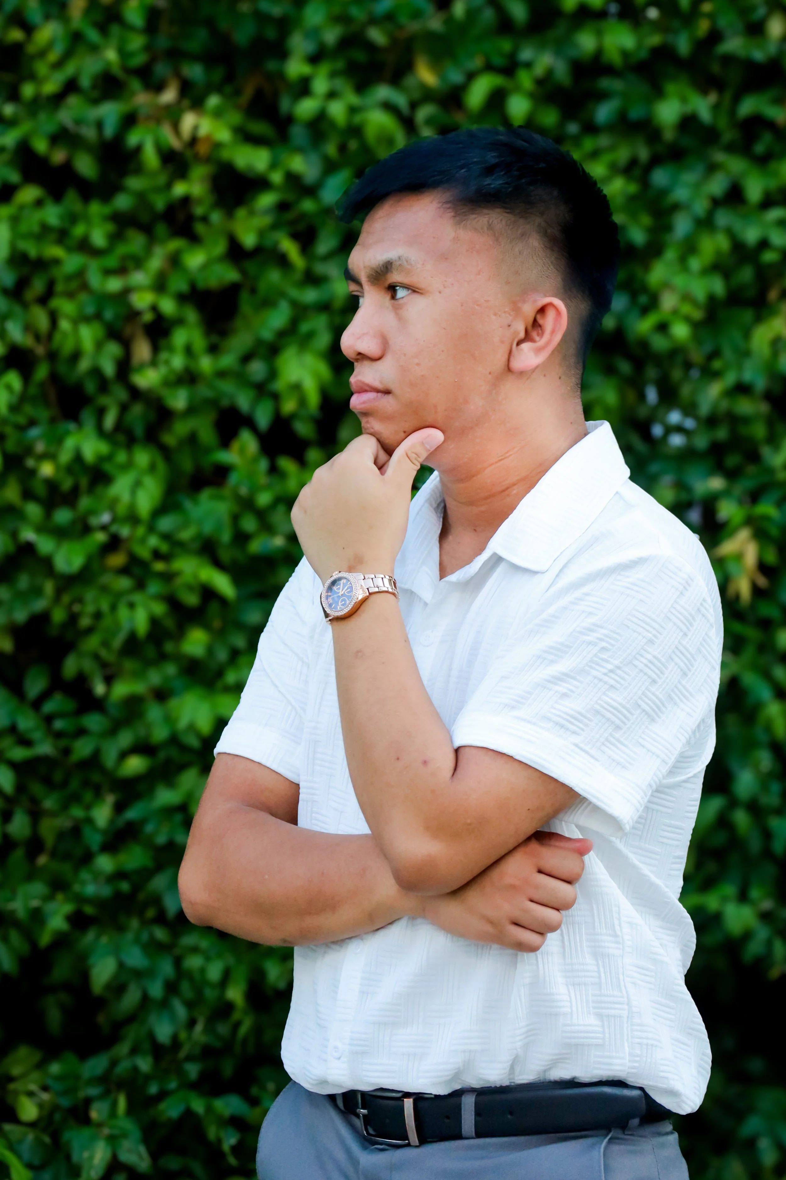 A young man with short black hair, wearing a white short-sleeved shirt, gray pants, and a silver watch, standing outdoors with green foliage in the background. He is standing in profile, with one hand resting on his chin and the other crossed over hi