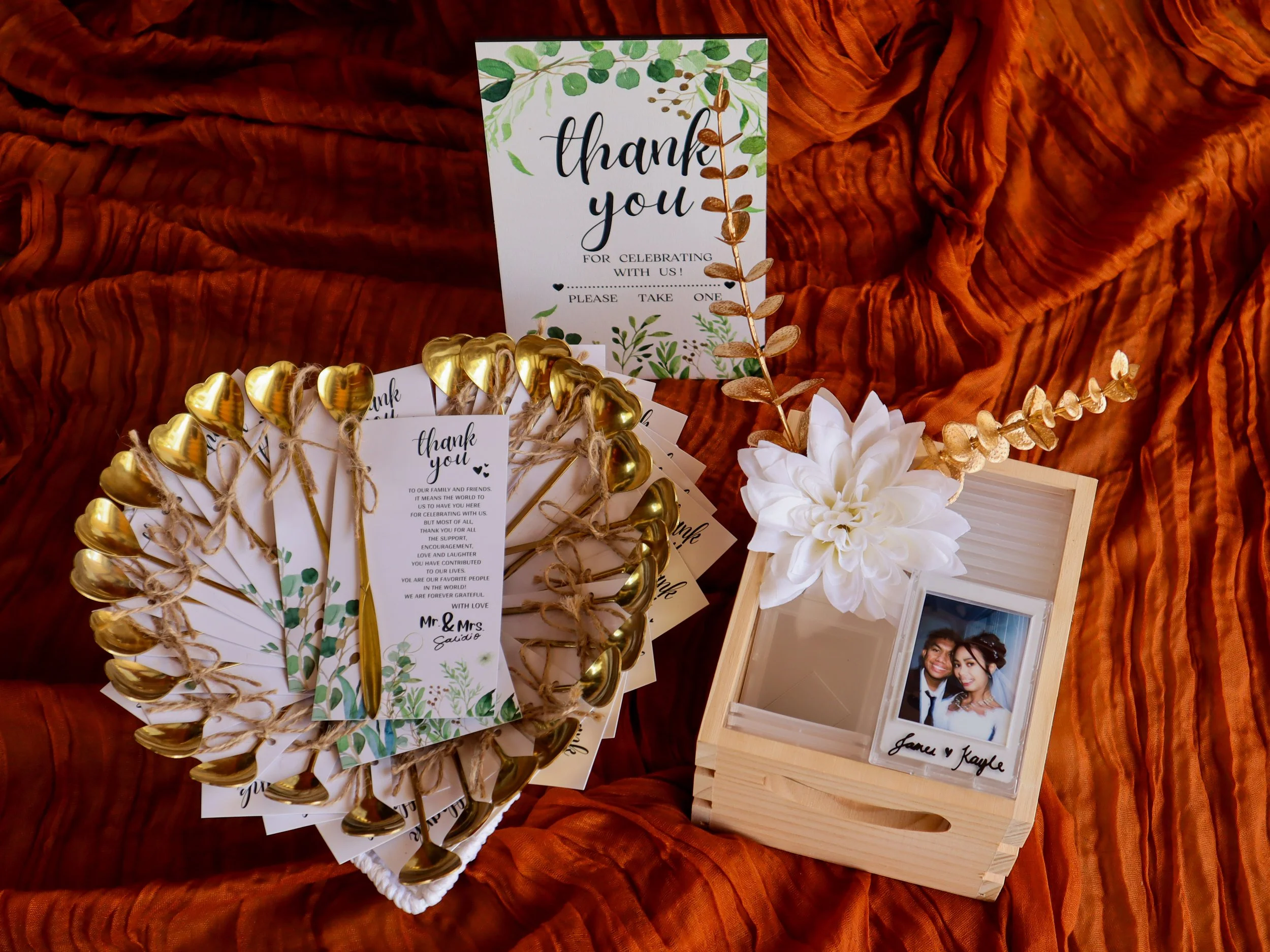 Wedding thank you cards, gold heart-shaped spoons, a framed photo of the couple, and a bouquet on a rust-colored textured fabric background.