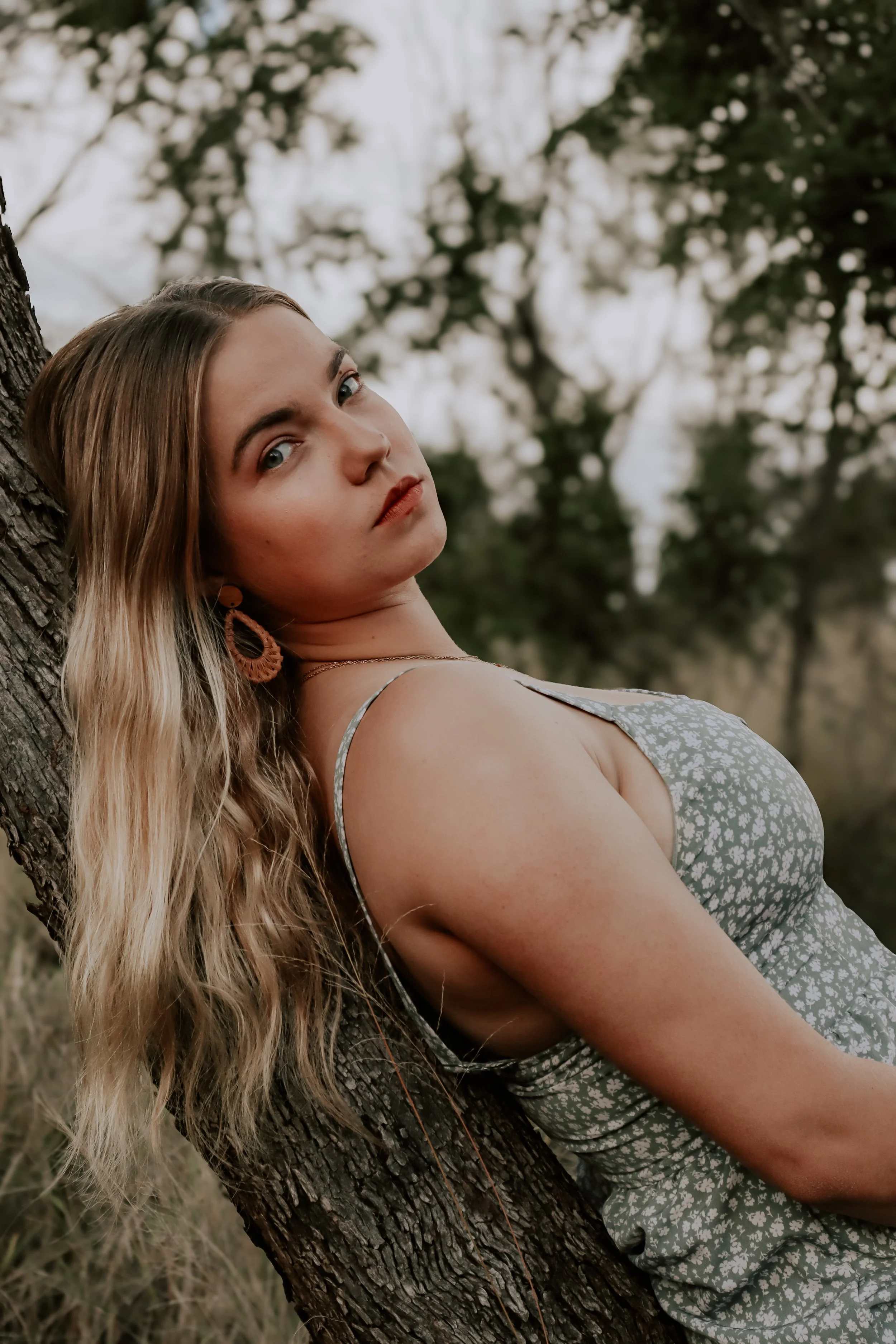 A woman with long blonde wavy hair leaning against a tree outdoors, wearing a light gray patterned dress and large earrings.