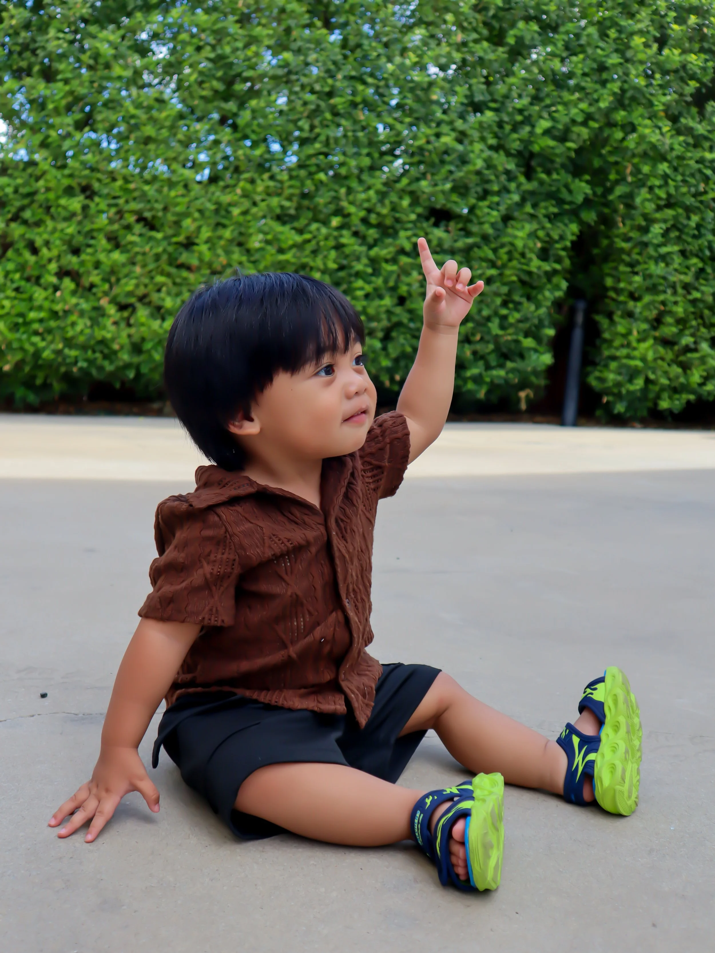 A young child with black hair sitting on concrete ground, pointing upward with his index finger. He is wearing a brown shirt, black shorts, and bright green and blue sandals. Background has green bushes.
