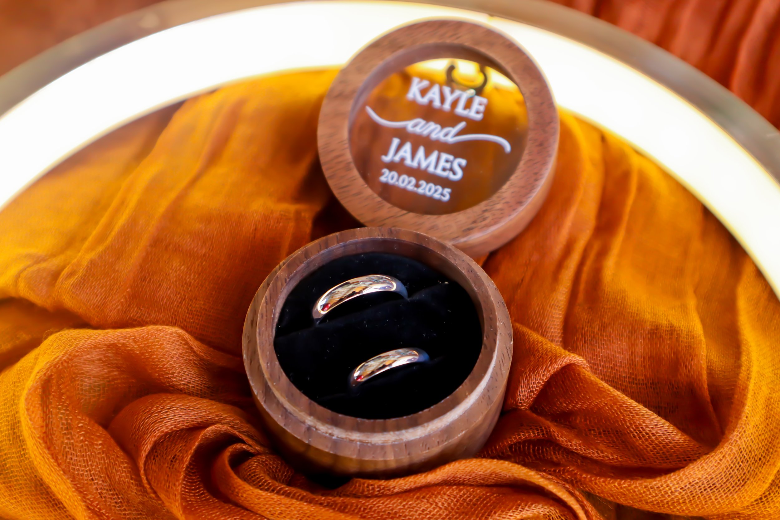A pair of wedding rings in a wooden box next to a round wooden sign engraved with names Kayle and James and the date 20.02.2025, all placed on an orange cloth.