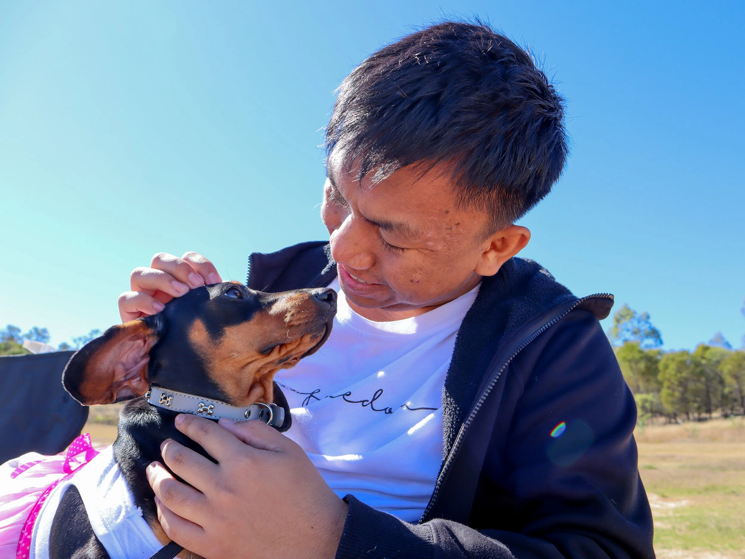 A man holding a small black and tan dog, looking into the dog's eyes outdoors on a sunny day.