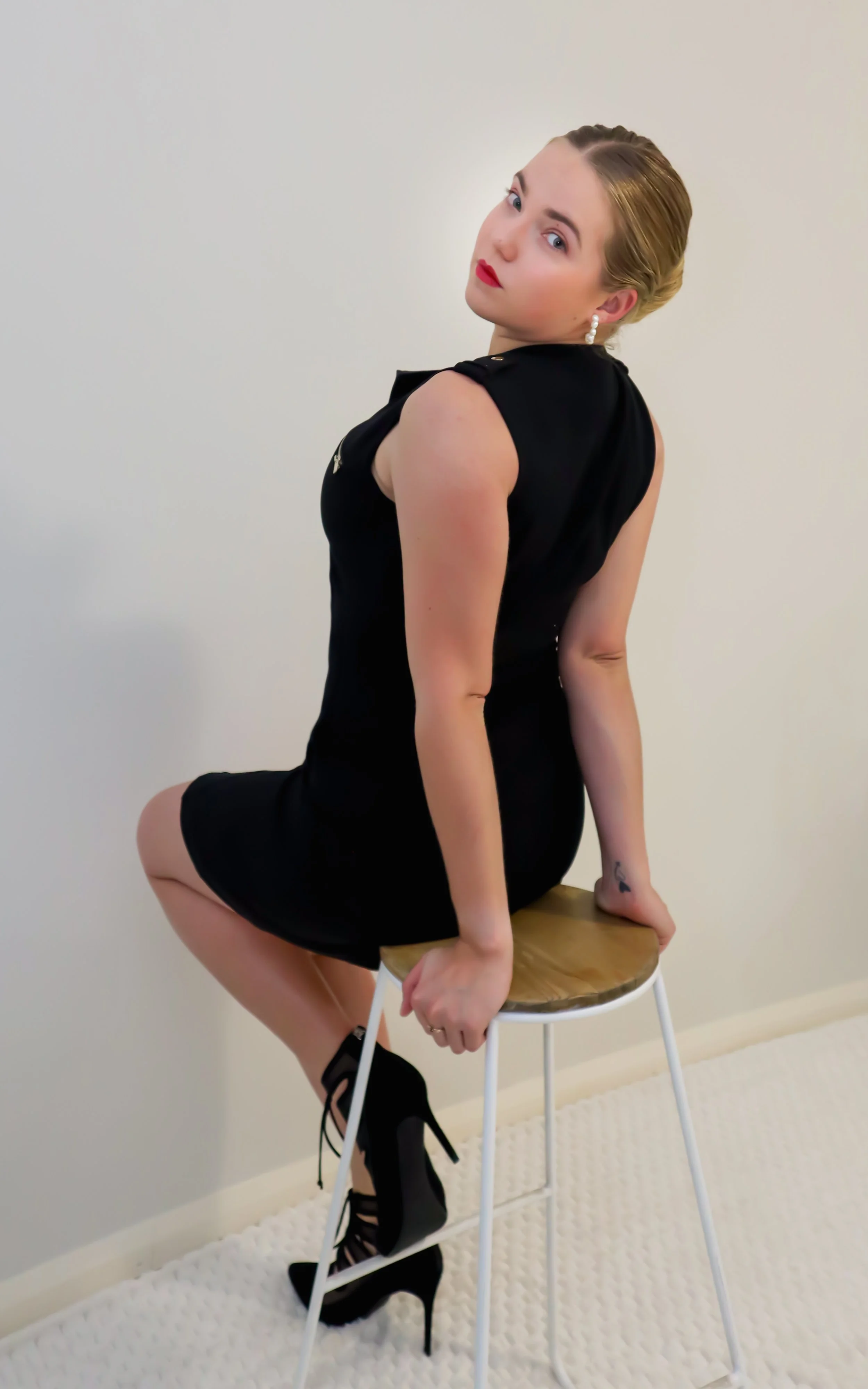 A woman with blonde hair styled back, wearing bright red lipstick, pearl earrings, a black sleeveless dress, and black high-heeled shoes, sitting sideways on a white stool with a round wooden seat, in front of a plain white wall.