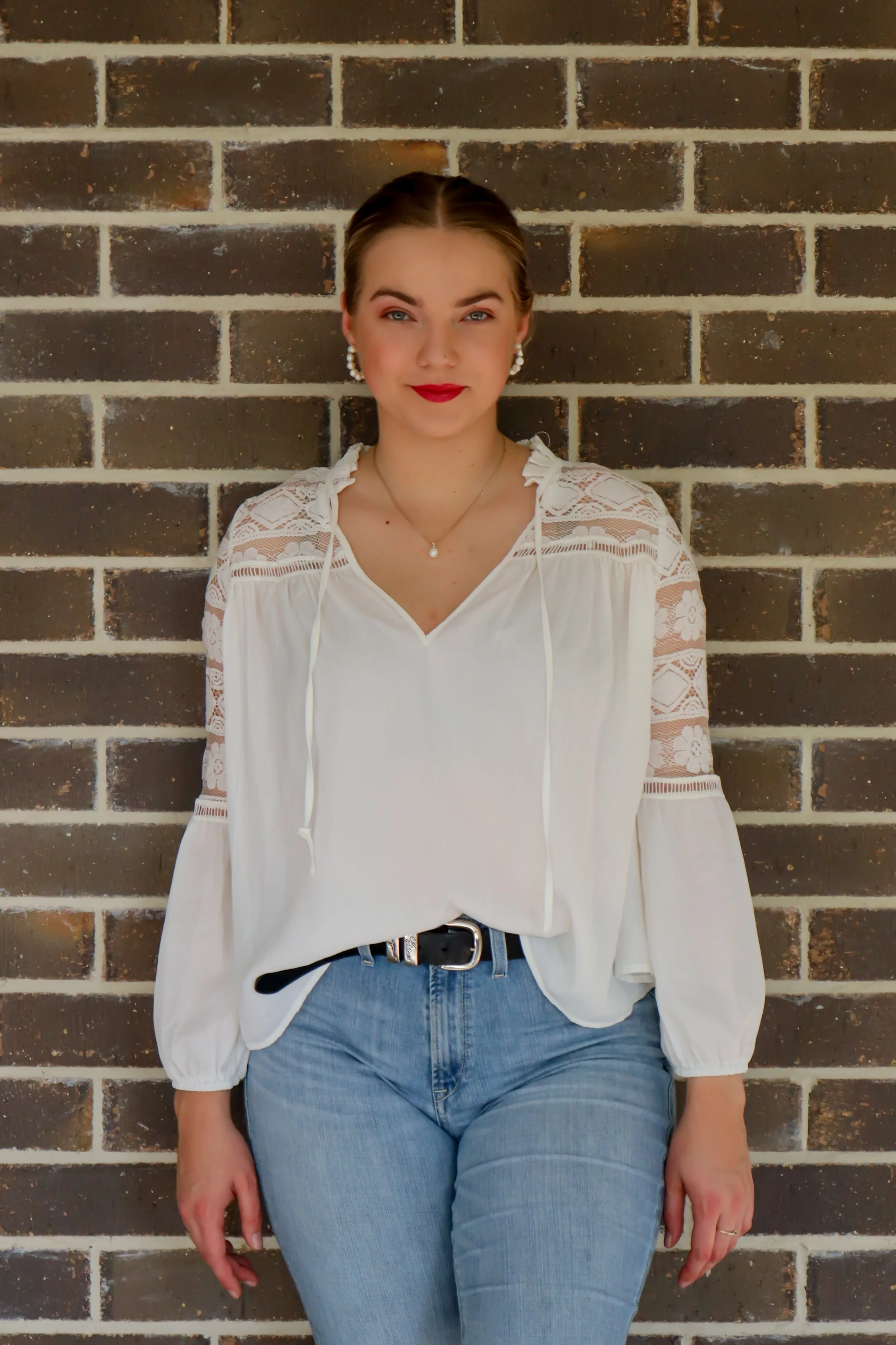 Young woman with blonde hair, wearing a white blouse with lace sleeves, blue jeans, and pearl earrings, standing against a brick wall.