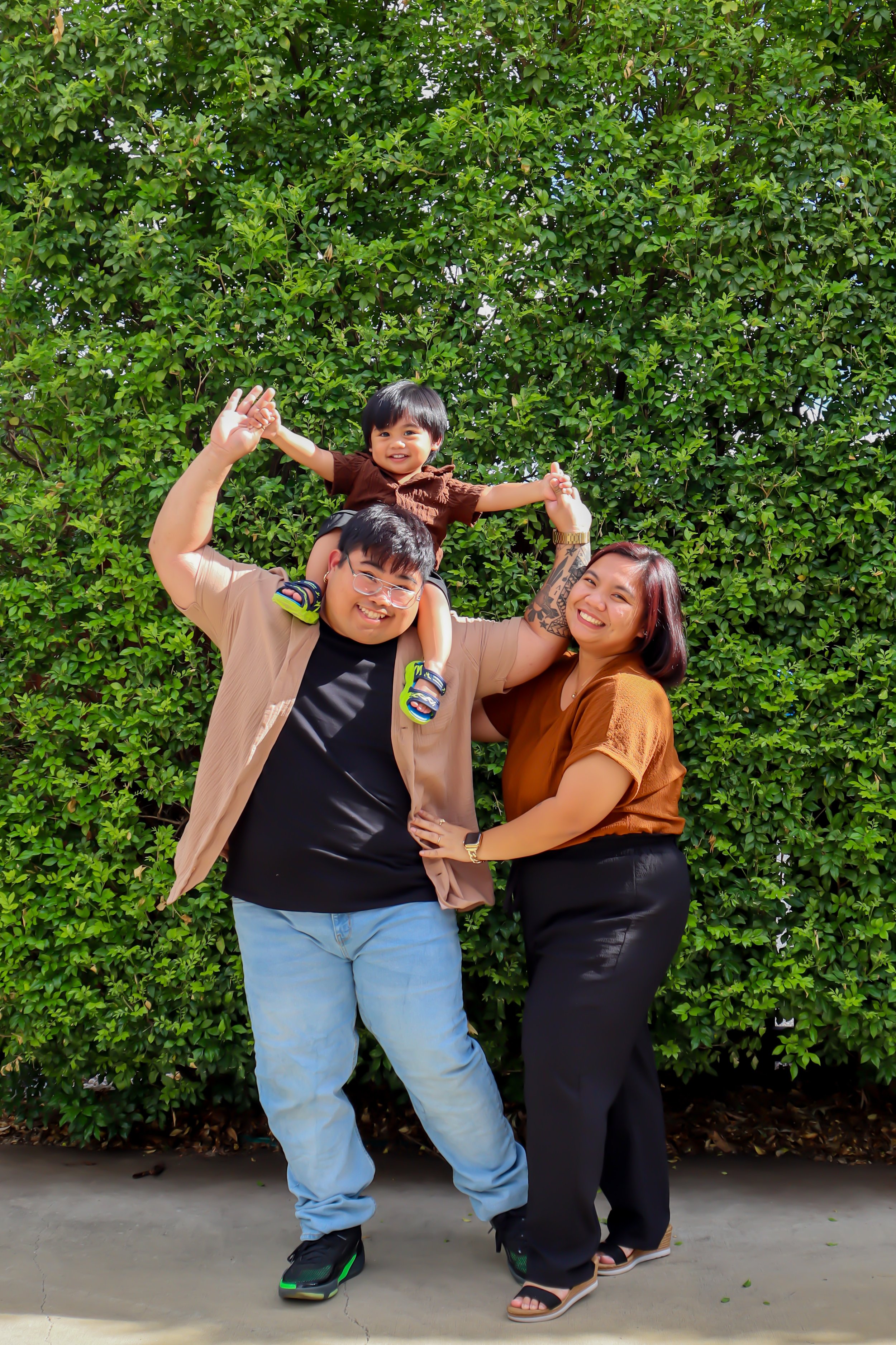 Family of three smiling and posing outdoors in front of lush green bushes. The father carries a young child on his shoulders, and the mother stands next to them, holding the father’s waist.