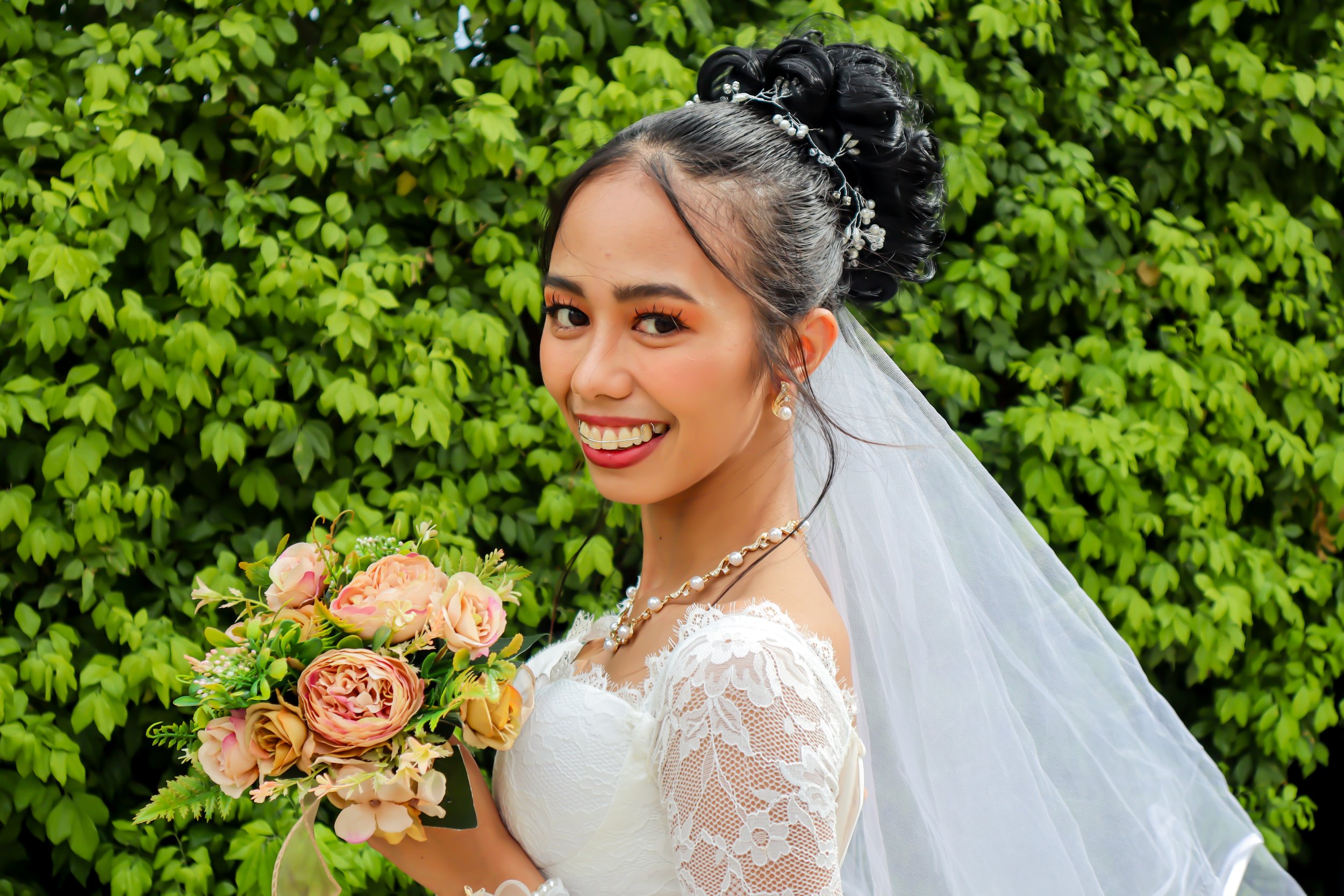 A smiling bride in a white lace wedding dress holding a bouquet of pink and peach flowers, standing outdoors in front of green foliage.