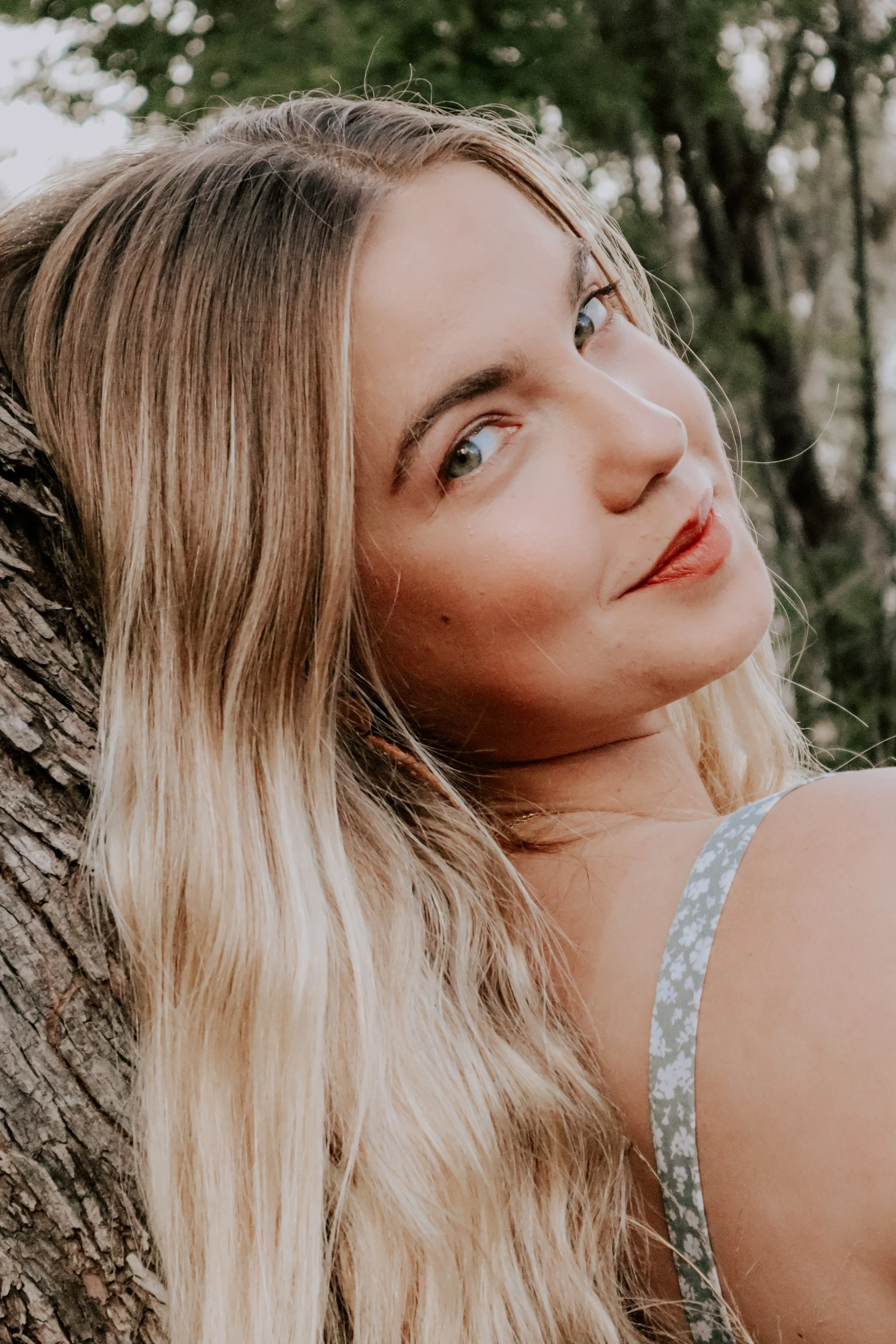 Close-up of a young woman with long blonde hair leaning against a tree outdoors, looking at the camera with a slight smile.