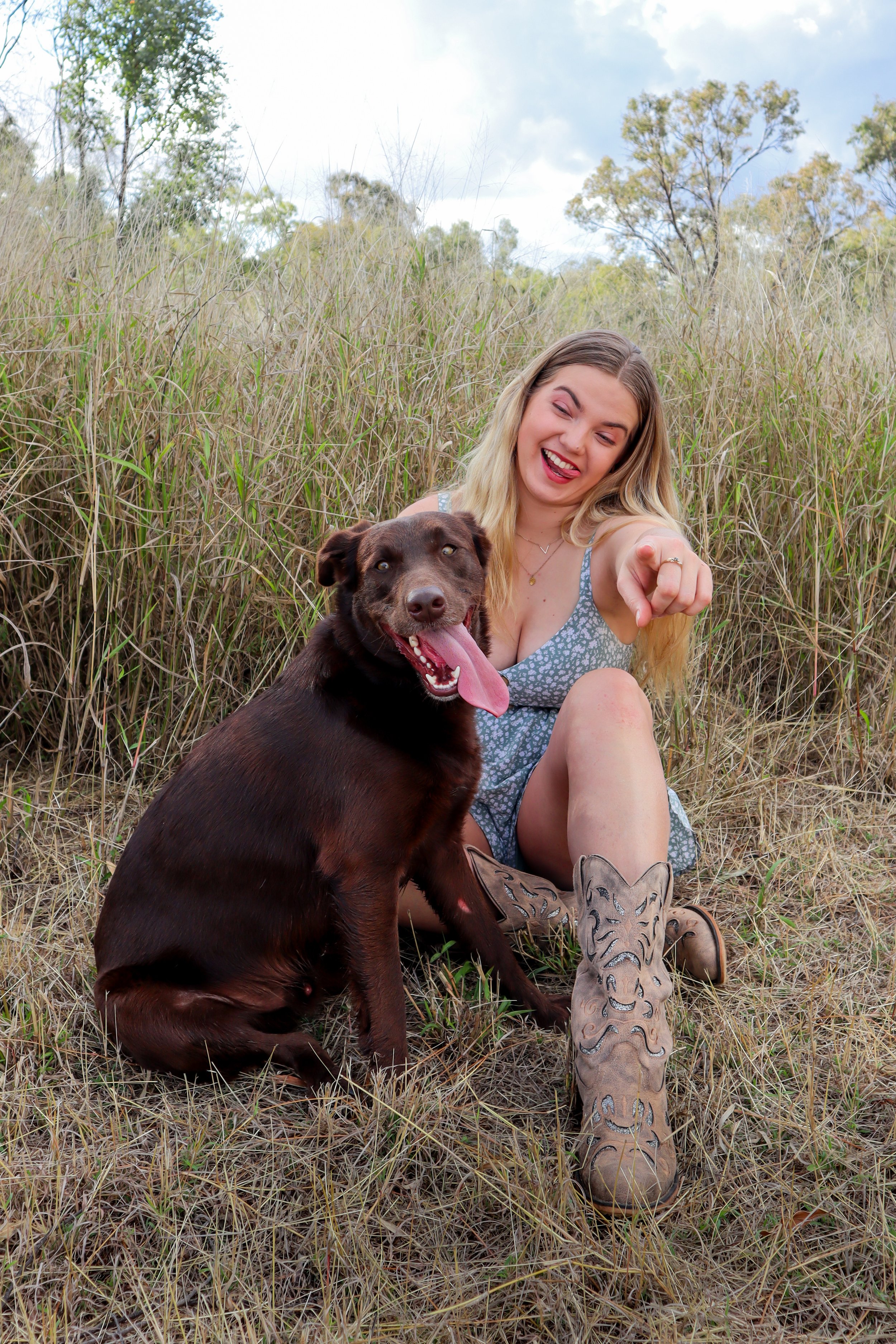 A young woman with blonde hair and a dog sitting in tall grass in a natural outdoor setting, the woman is laughing and pointing, wearing a blue patterned dress and cowboy boots with a tiger design.