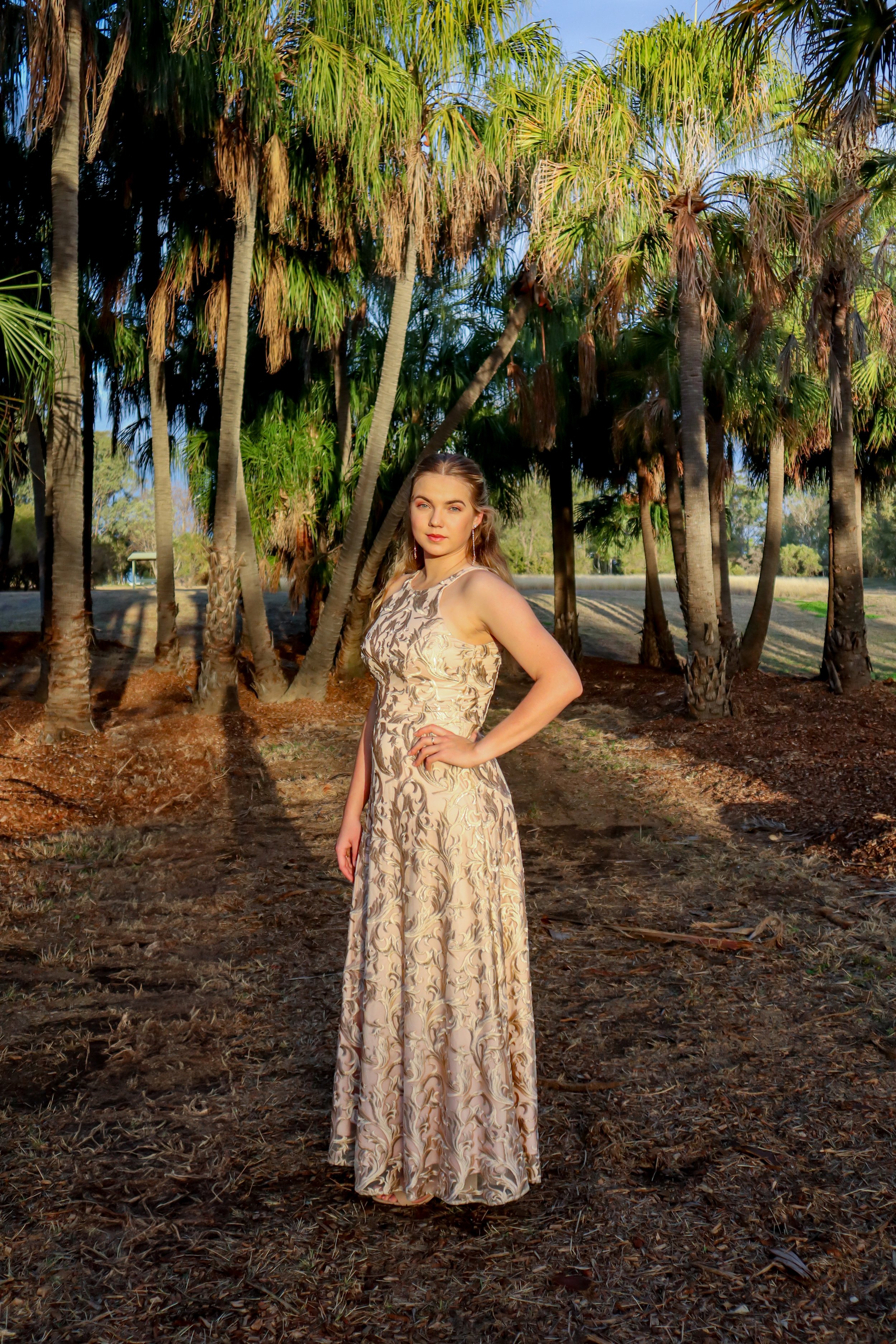 A young woman in a patterned, sleeveless, long dress standing on a dirt path among tall palm trees in a sunny outdoor setting.