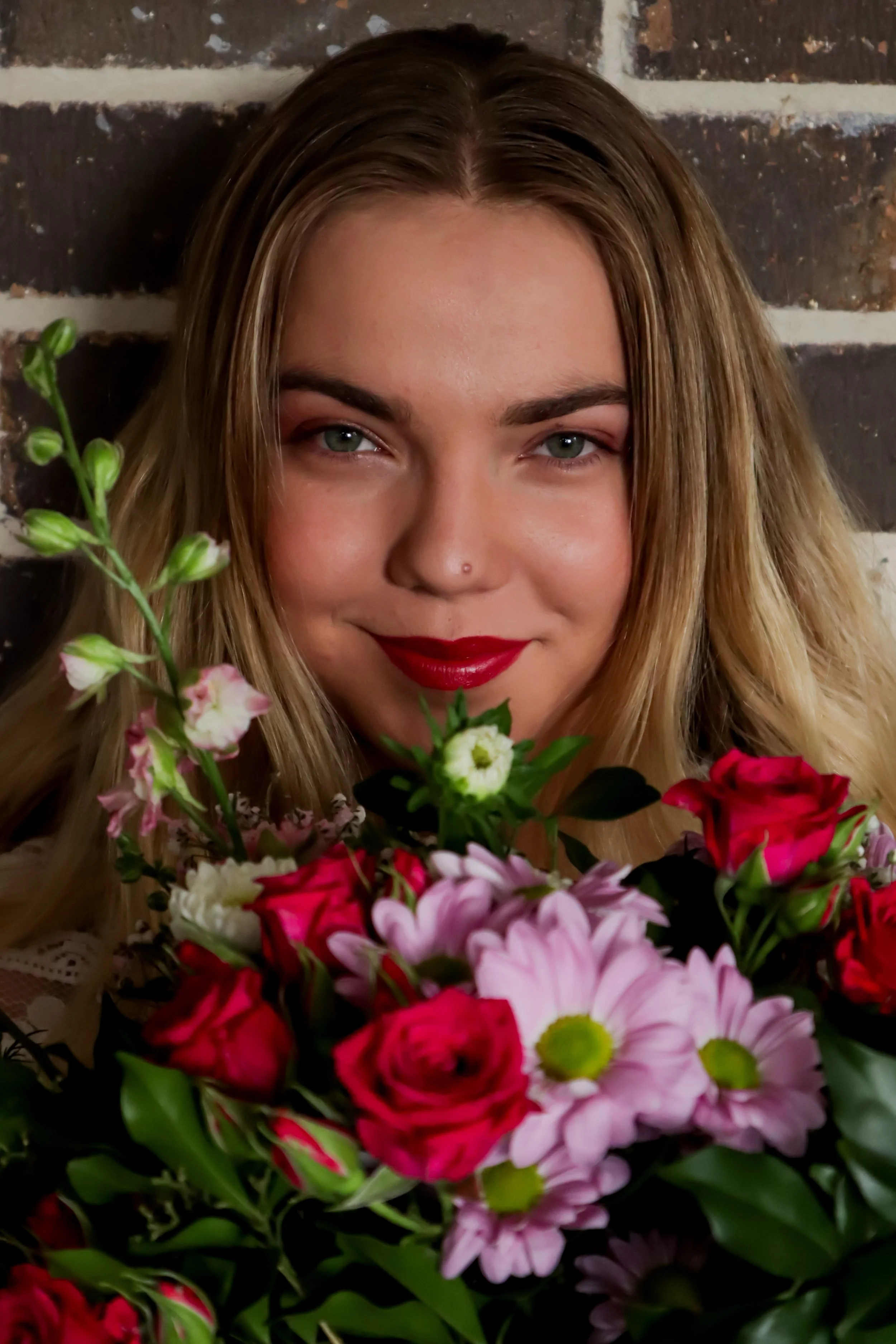A woman with blonde hair, red lipstick, and a nose piercing holds a bouquet of pink, red, and white flowers with a brick wall background.