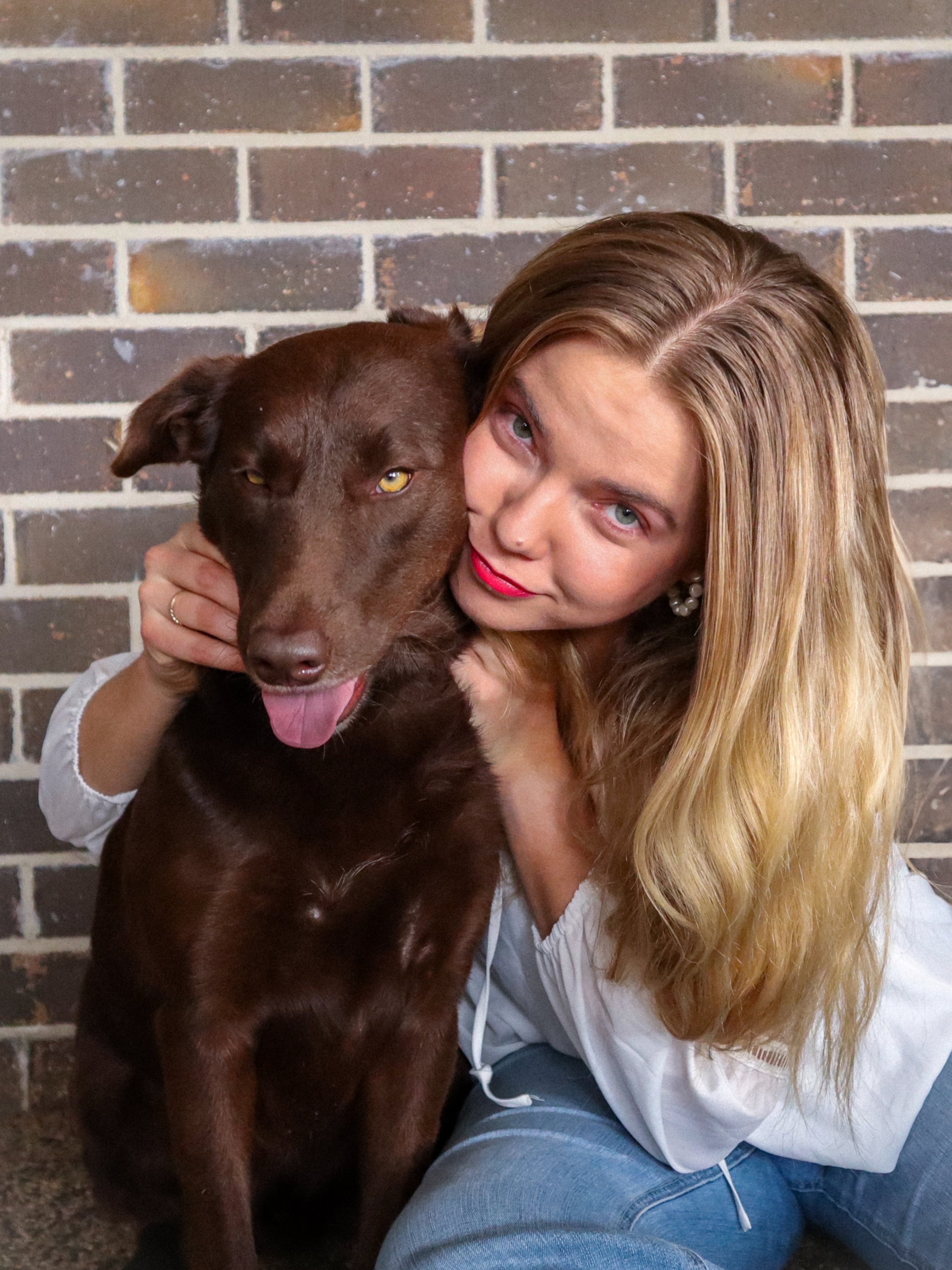 A woman with long blonde hair, wearing a white top and red lipstick, holding a brown dog with yellow eyes against a brick wall background.