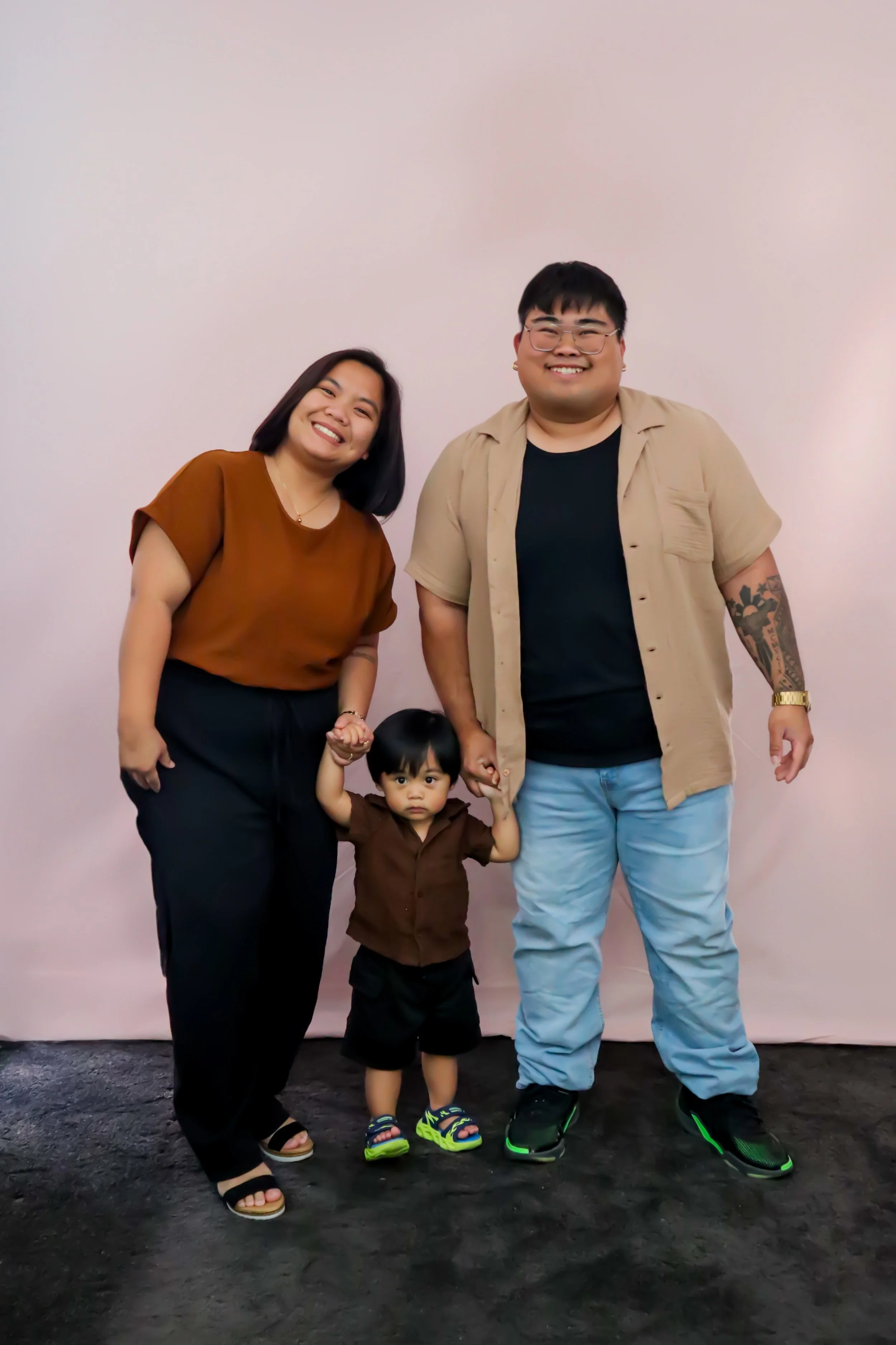 A family of three holding hands and smiling at the camera against a pink backdrop. The woman and man are adults, and there is a young child between them.