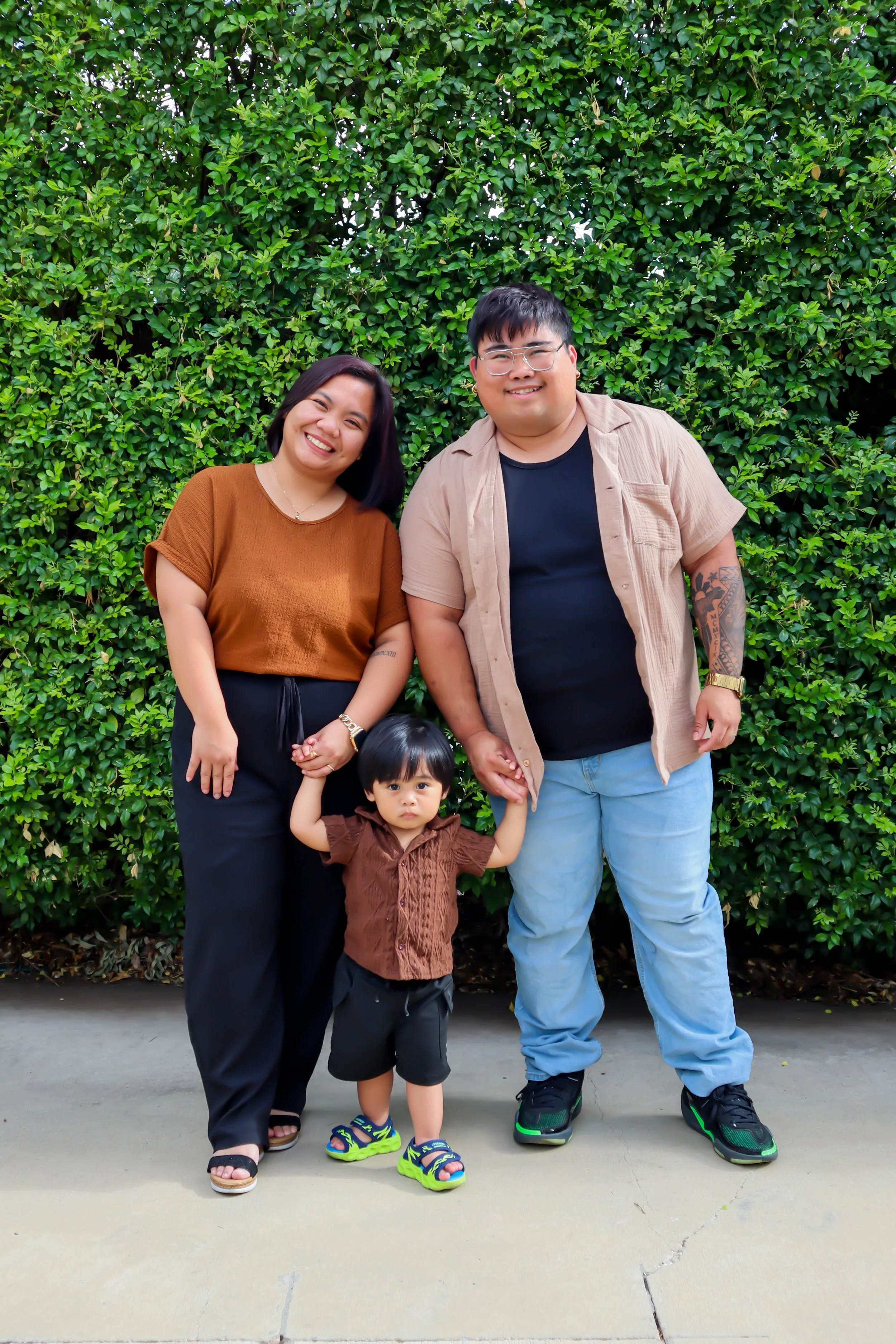 A family of three smiling and holding hands in front of a green hedge. The woman has shoulder-length dark hair and is wearing a brown top and black pants. The man has short dark hair, glasses, and is wearing a beige short-sleeved button-up shirt over