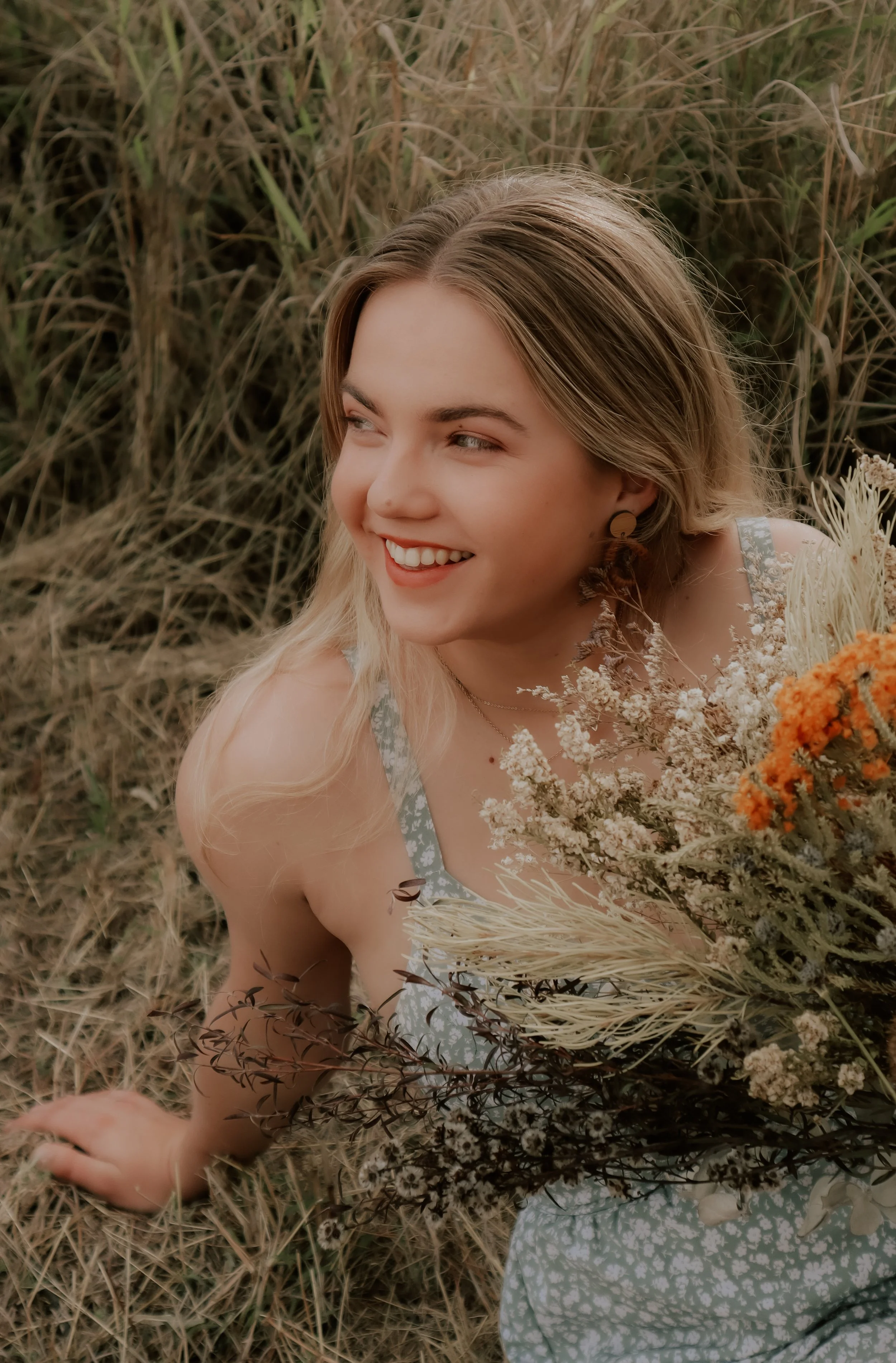 A young woman with blonde hair and a light floral dress sitting on grass among dry plants, holding a bouquet of wildflowers, smiling and looking to her left.