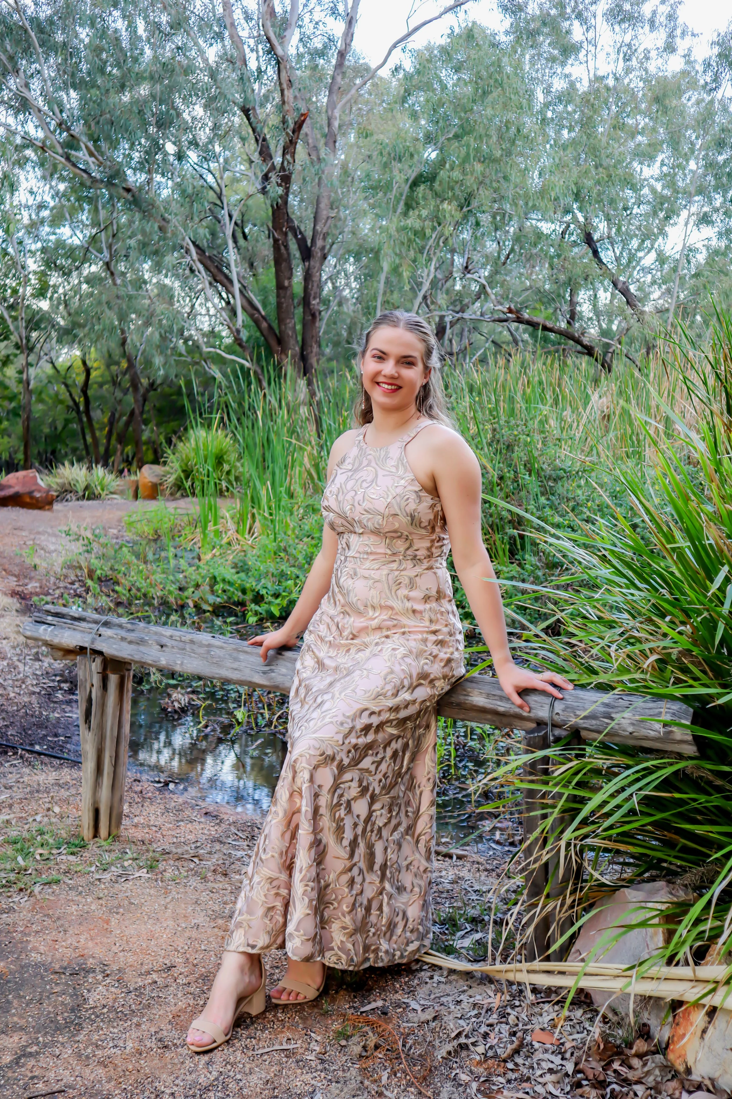 A young woman in a long, patterned dress sitting on a wooden bench outdoors surrounded by greenery and trees, smiling at the camera.