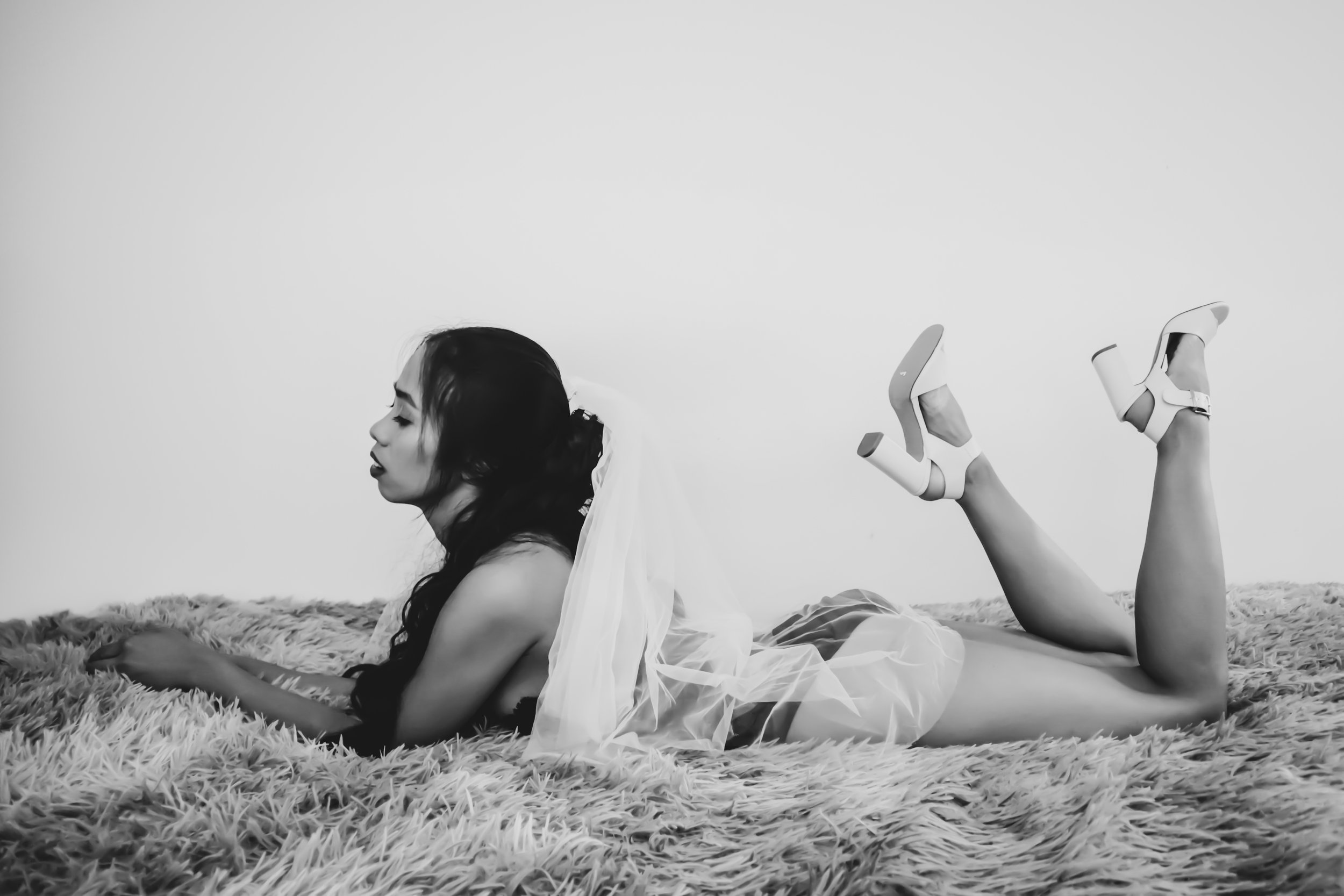 A woman in high heels and a veil, lying on her stomach on a plush rug, with her legs bent at the knees and slightly raised, holding a phone in her hand, in a black and white photo.