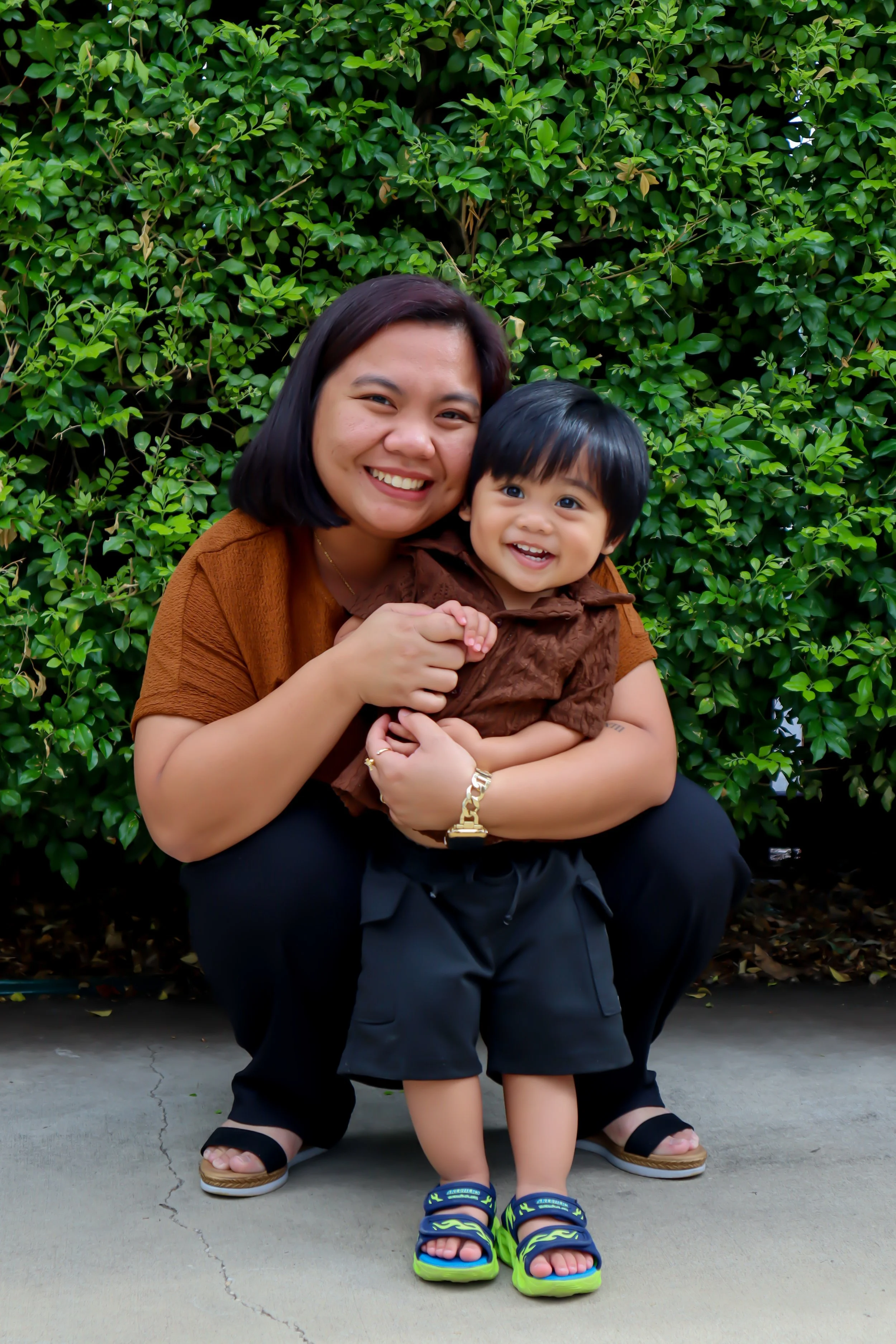 A woman and a young boy smiling and hugging outdoors in front of a leafy green bush.