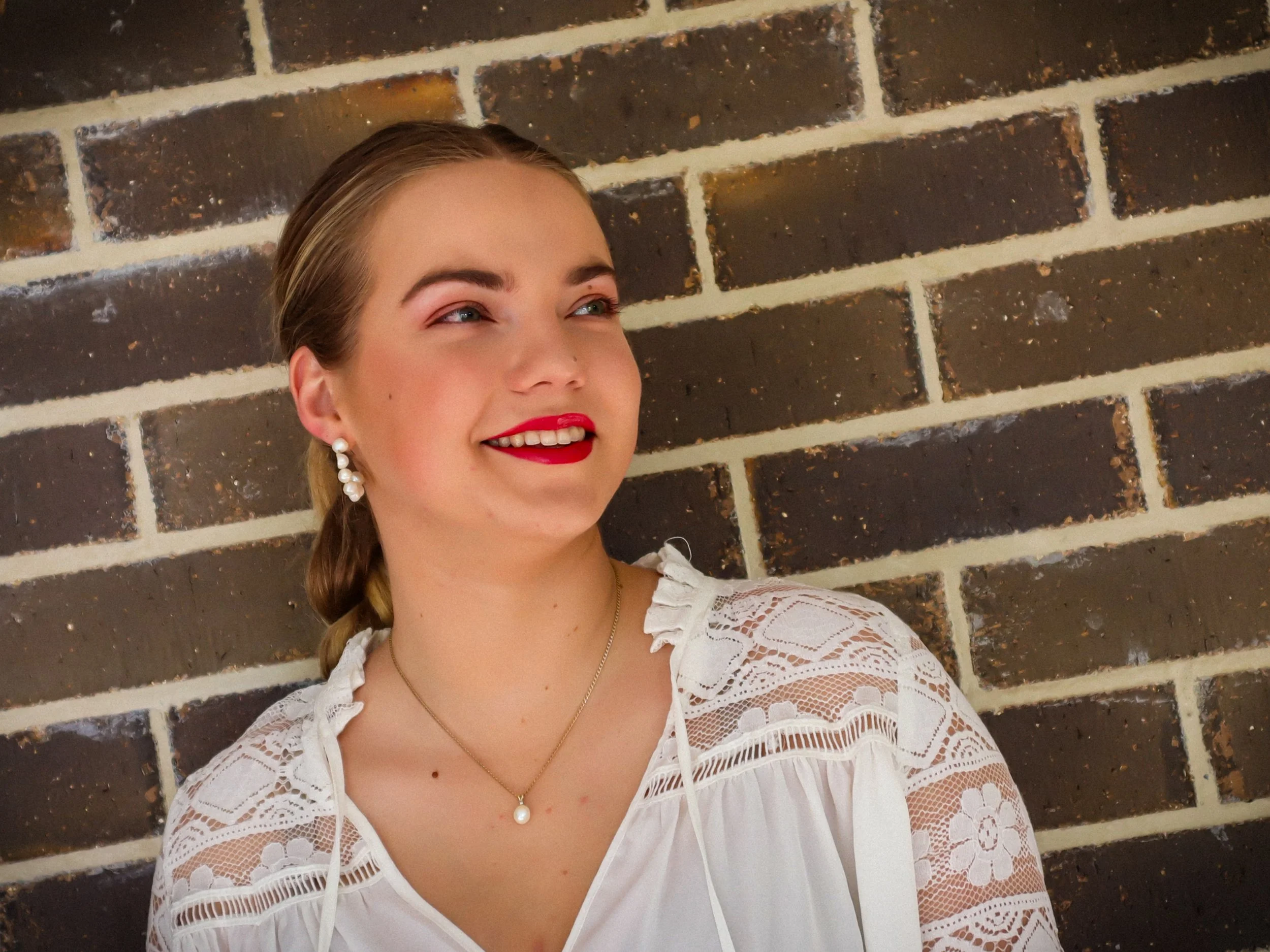 A young woman with blonde hair, wearing a white lace blouse, pearl earrings, and a pearl necklace, smiling and looking to the side against a brick wall background.