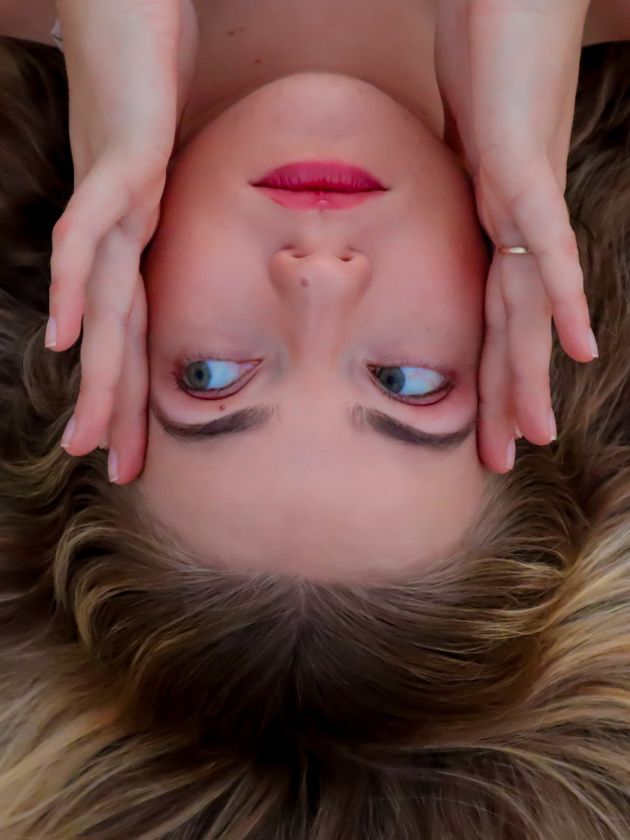 A woman with light skin and brown hair lying down with her eyes open, resting her head on a furry surface, with her hands framing her face.