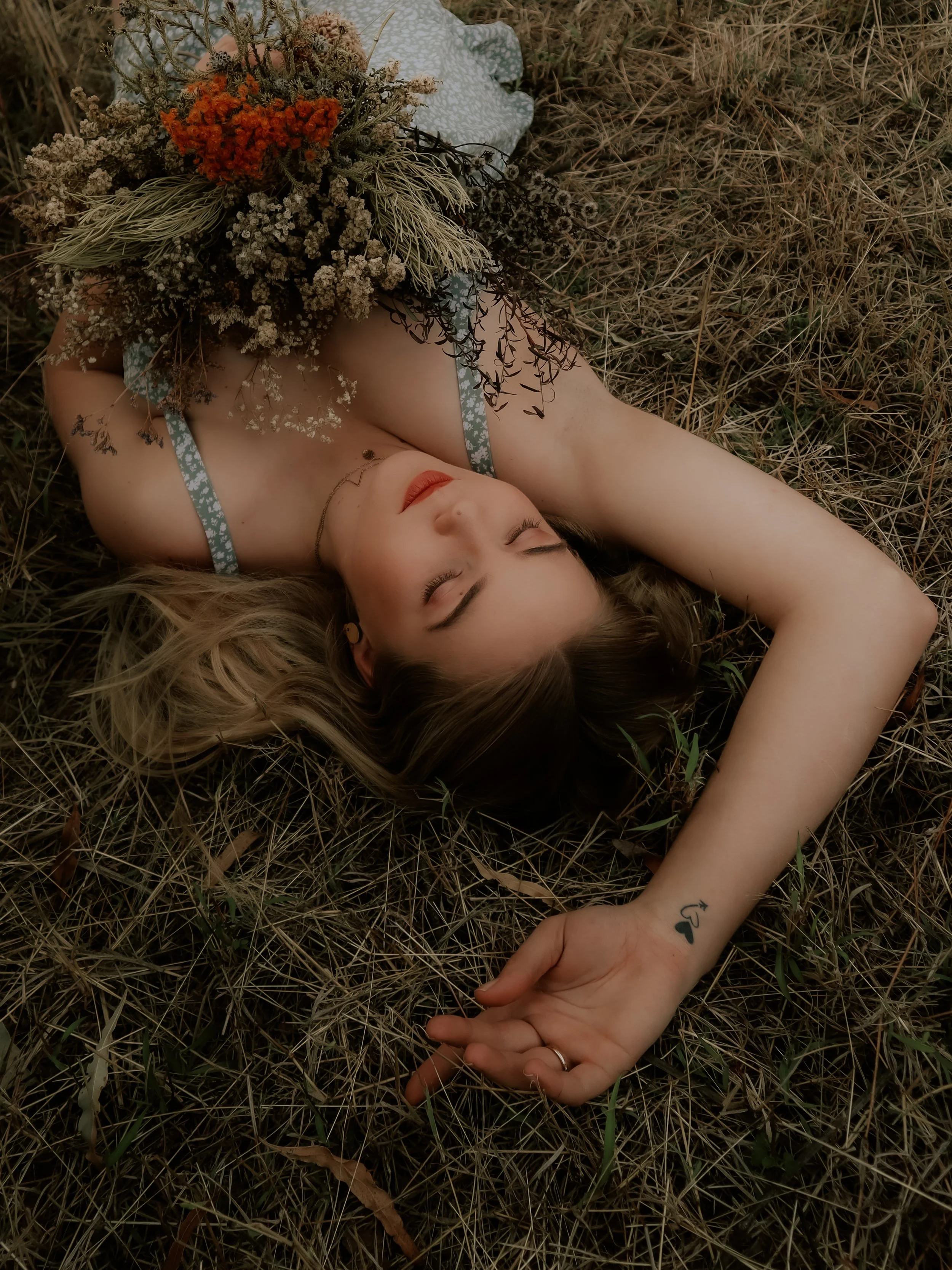 A young woman lies on her back on dry grass with her eyes closed, holding a bouquet of dried flowers on her chest. She has blonde hair, red lipstick, and a small tattoo of a heart with an arrow on her left wrist. She is wearing a blue and white flora