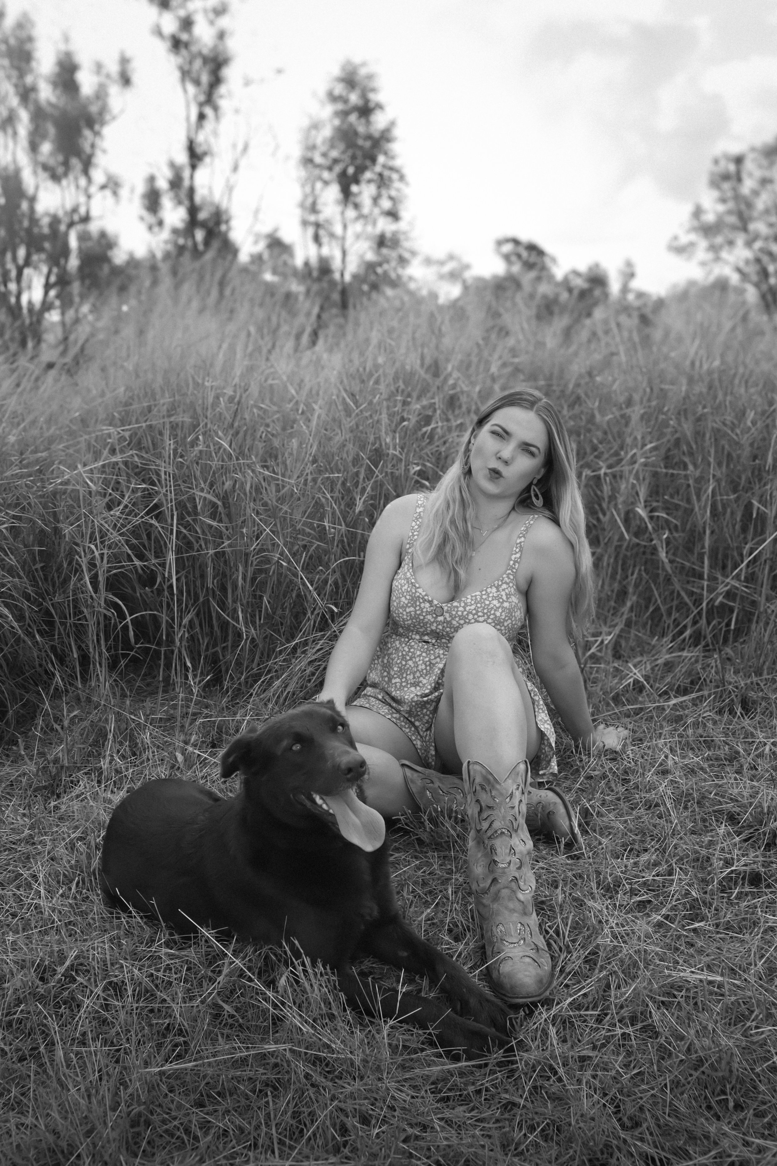 A woman sitting on grass in a field with tall grass and trees in the background, posing with a dog and a carved wooden mask.
