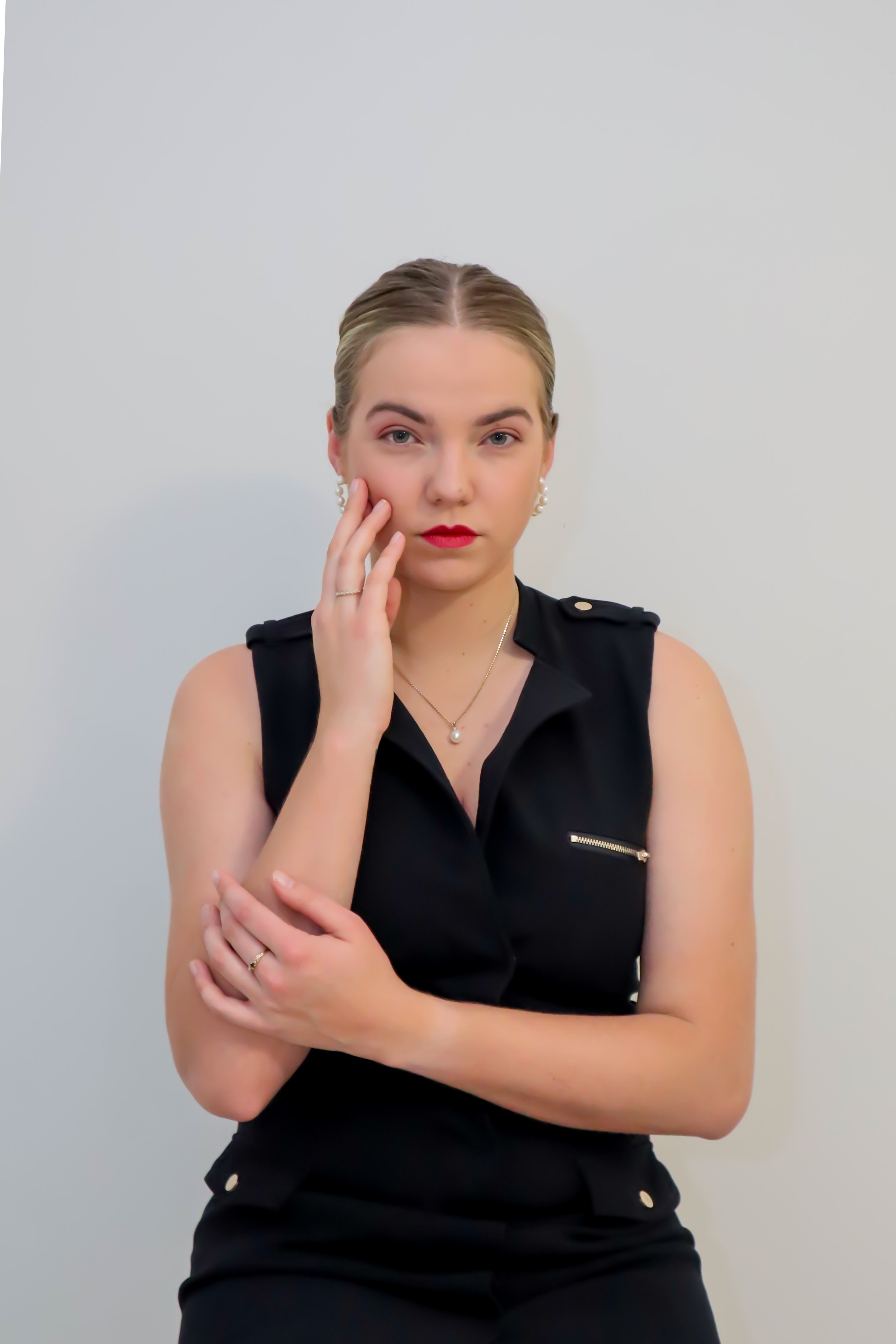 A young woman in a black sleeveless top stands against a plain white wall, touching her face with her left hand and crossing her arms over her chest. She has blonde hair, red lipstick, and jewelry including earrings, a necklace, and rings.