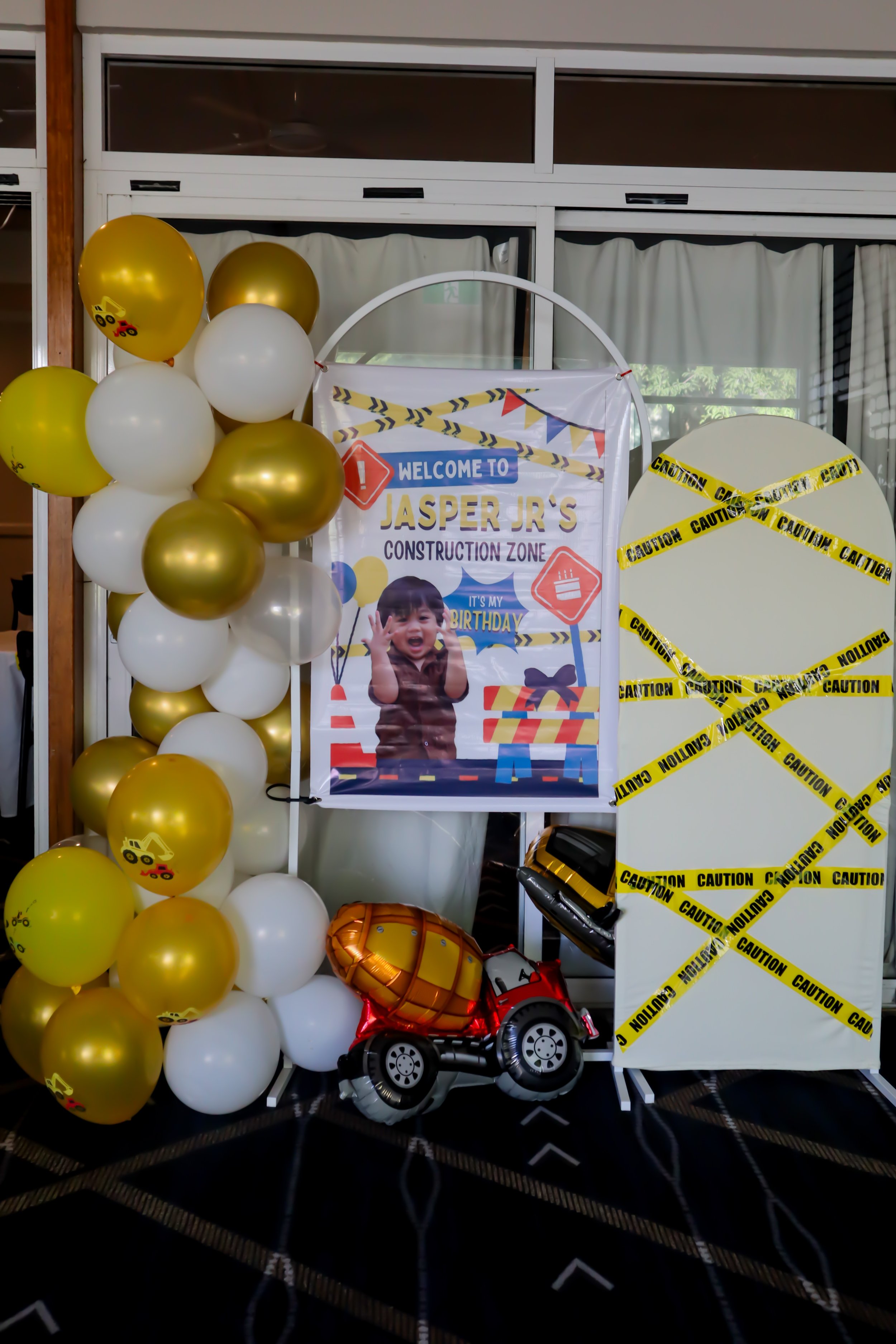 Construction-themed birthday party display with yellow and white balloons, a sign that reads 'Welcome to Jasper Jr.'s Construction Zone' featuring a smiling child, and caution tape decorations.