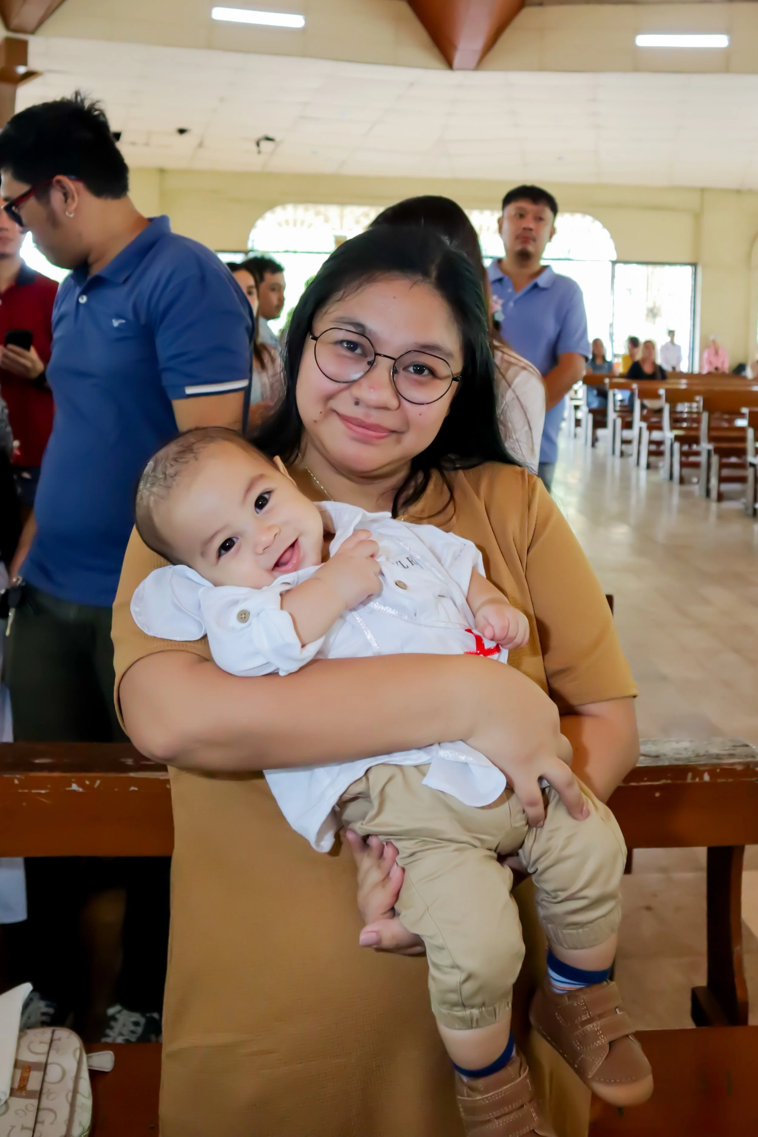 A woman holding a smiling baby in a church or chapel with other people in the background.