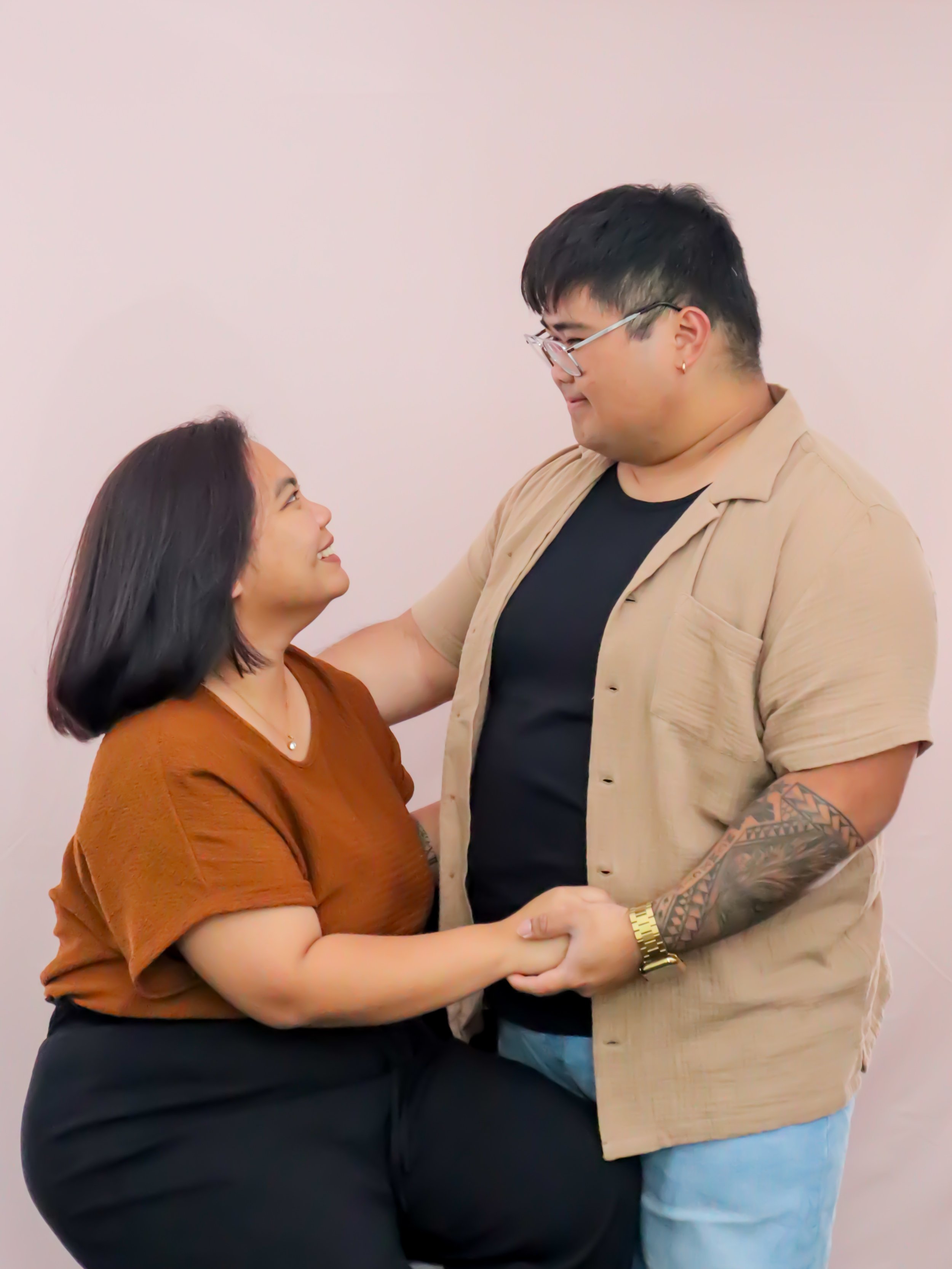 A couple holding hands and looking at each other with smiles, standing against a plain wall.