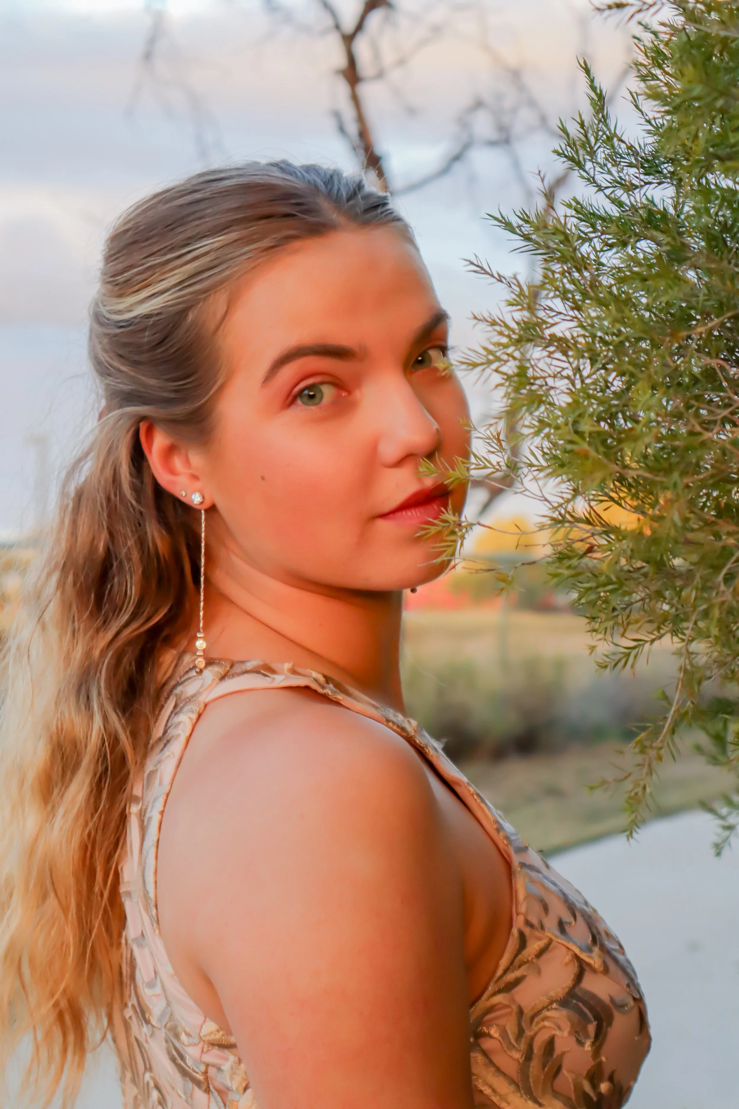 A young woman with long, wavy hair and earrings stands outdoors during sunset, partially obscured by a green bush, looking at the camera.