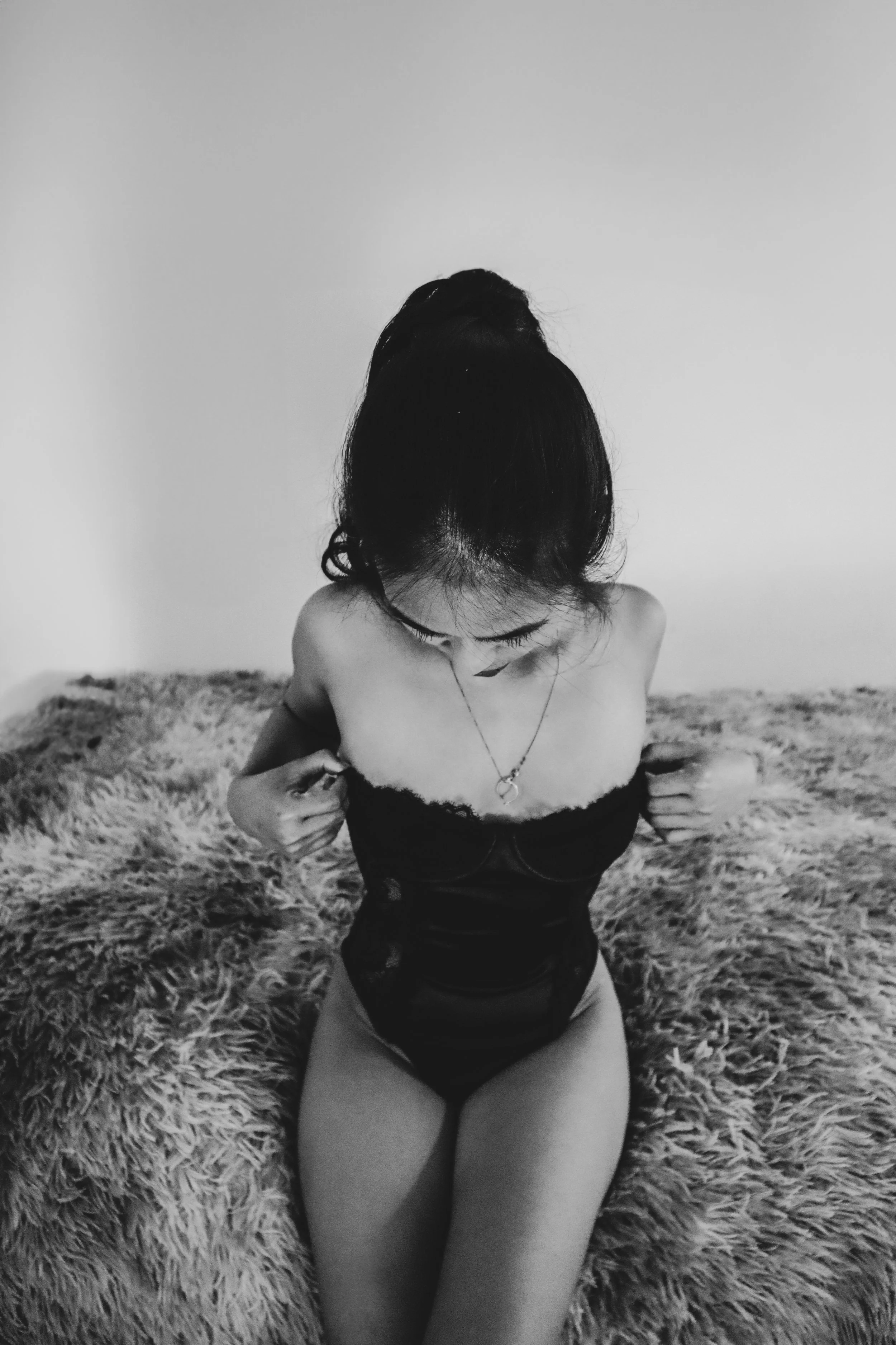 A woman with dark hair tied in a bun, wearing a strapless black top and a necklace, is sitting on a furry blanket, looking downward, with her hands adjusting her top.