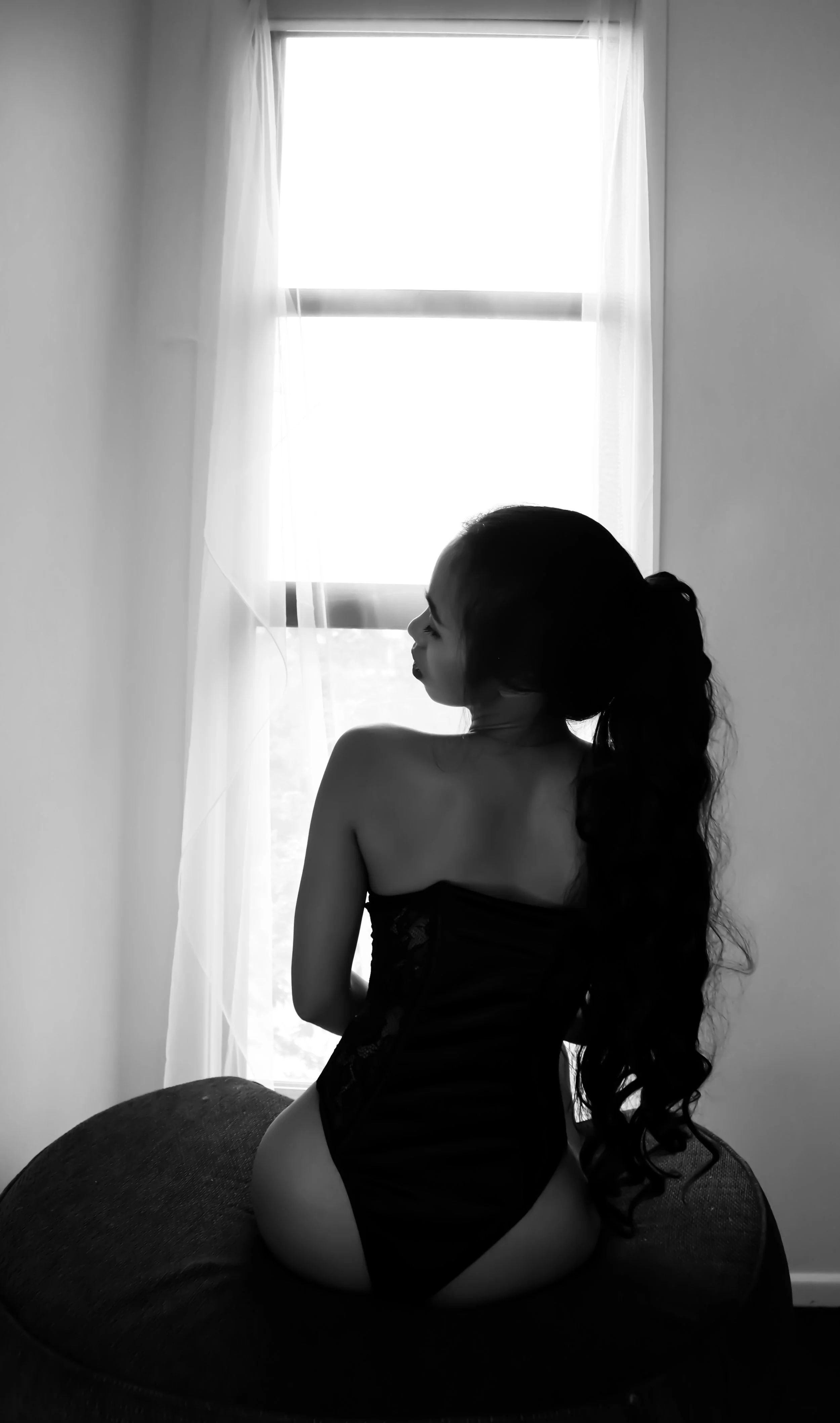 Black and white photo of a woman with long, curly hair sitting on a round cushion, facing away from the camera toward a window with light filters and sheer curtains.