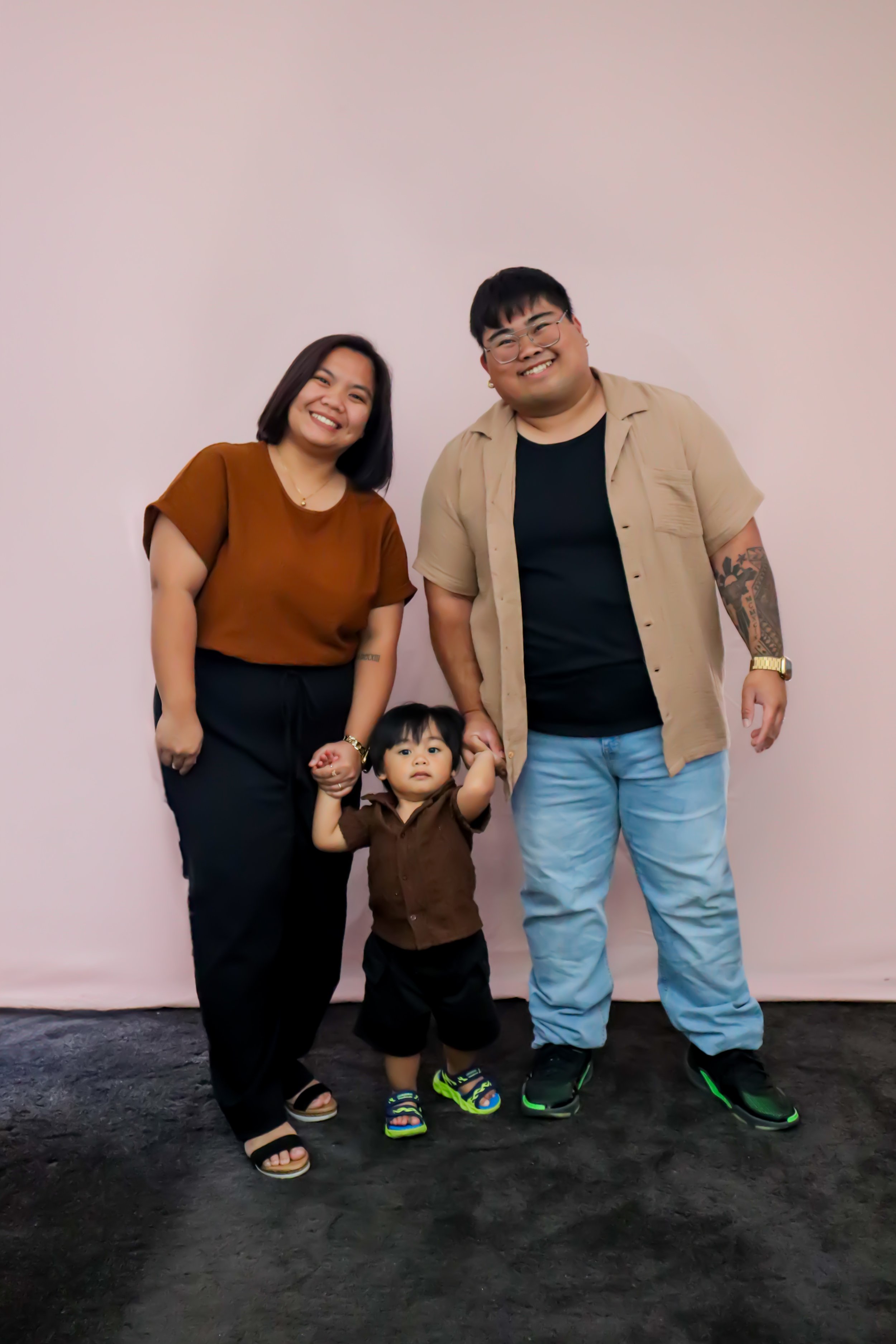 Family of three, a woman, a man and a young boy, standing together in front of a plain light-colored wall. The woman has shoulder-length dark hair, wearing a brown top and black pants. The man has glasses, short dark hair, wearing a beige short-sleev