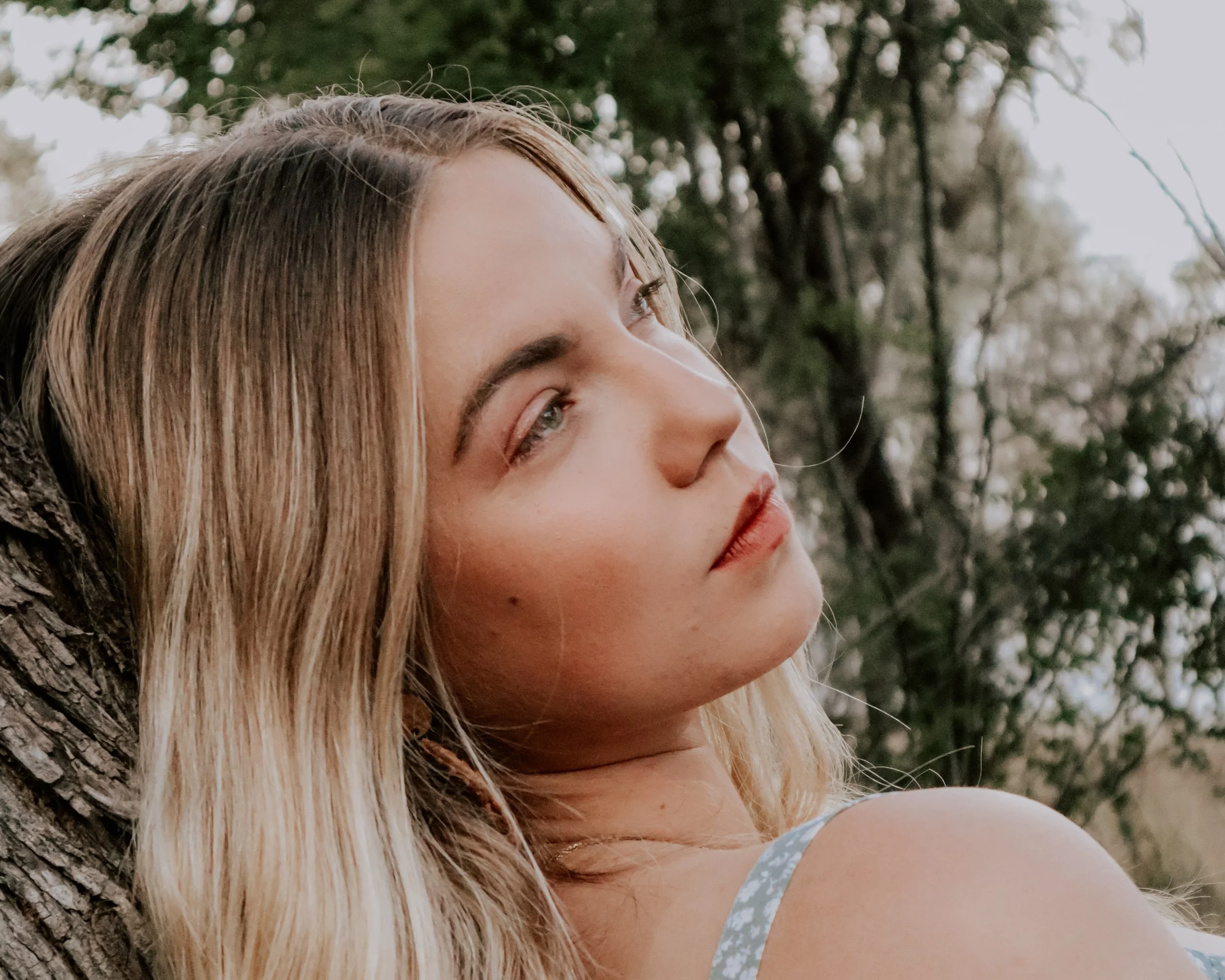 A young woman with blonde hair and light makeup leaning against a tree outdoors, looking pensively to the right, with a background of blurred trees.