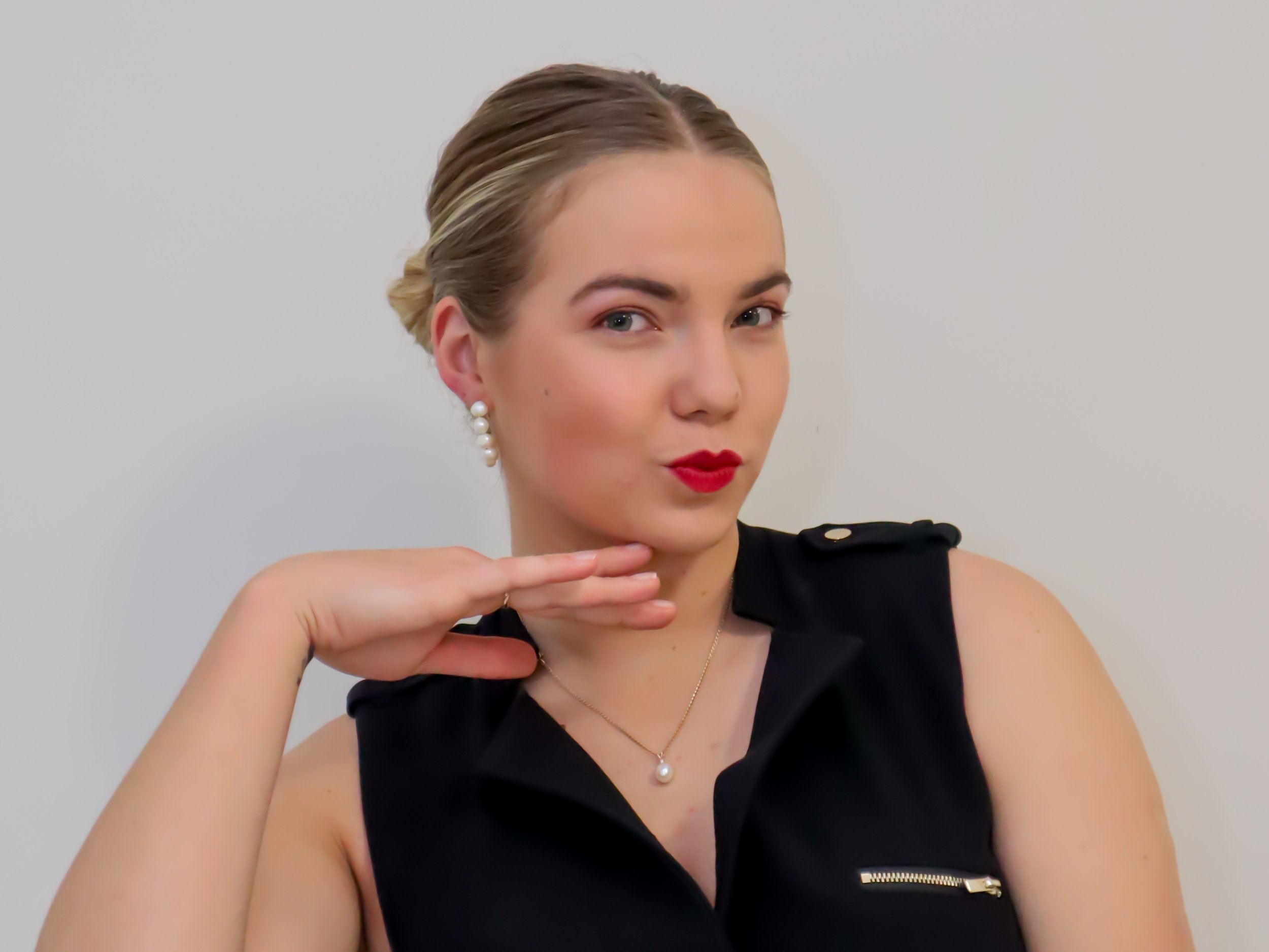 A woman with styled hair, red lipstick, and pearl earrings, wearing a black sleeveless top, striking a playful pose with her hand near her chin.