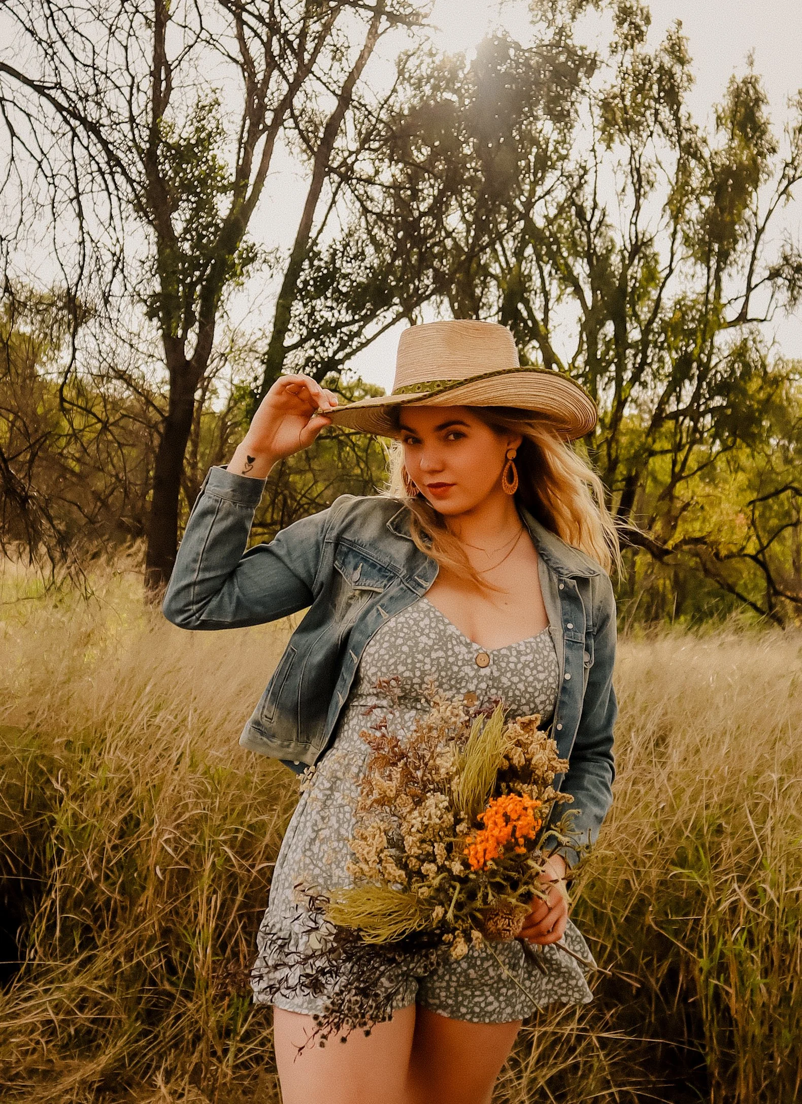 A young woman wearing a light-colored dress, denim jacket, and wide-brimmed straw hat stands in a field of tall grass, holding a bouquet of dried flowers, with trees and the setting sun in the background.