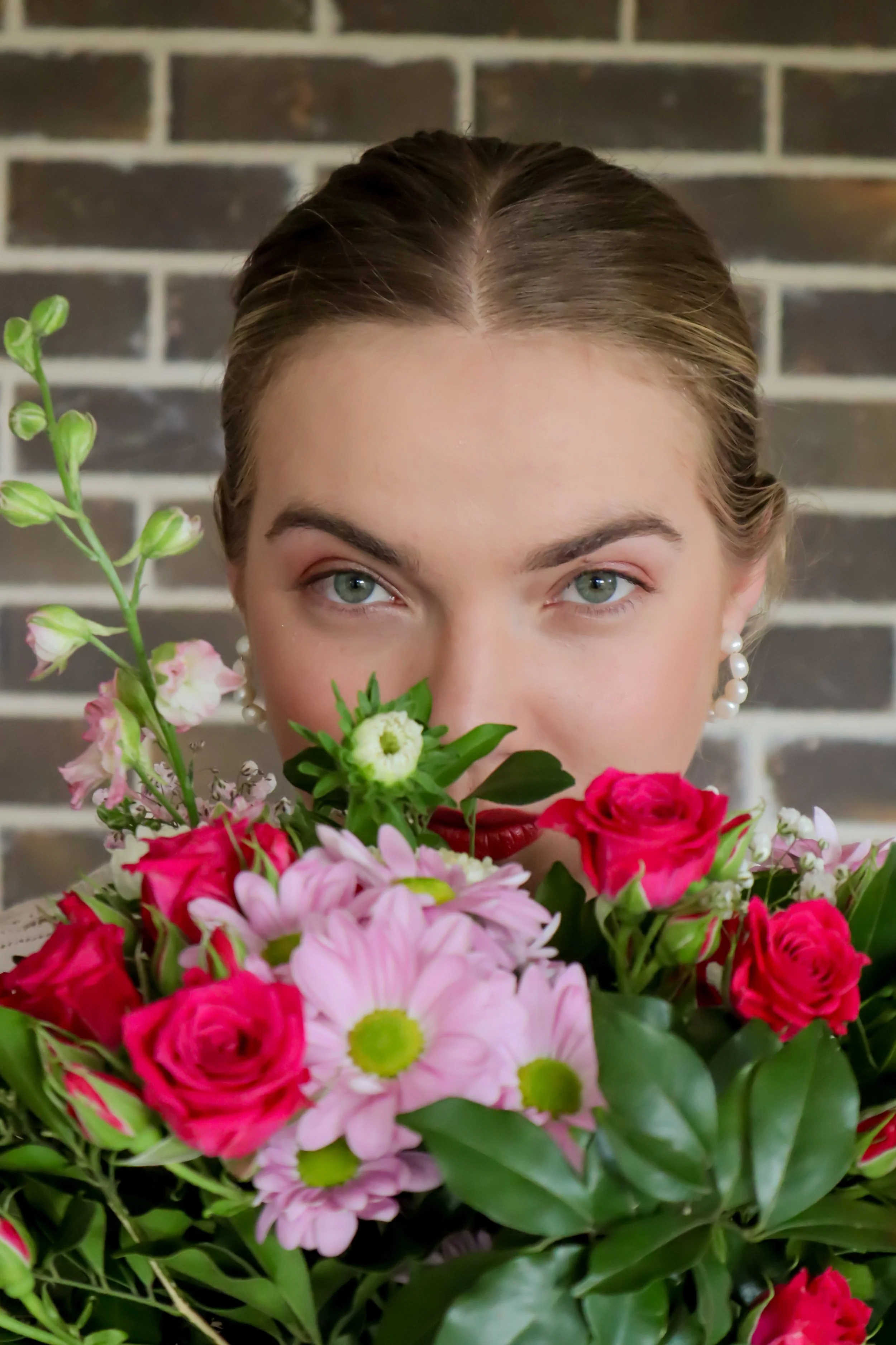 A young woman with pearl earrings holding a colorful bouquet of pink, red, and white flowers in front of her face, with only her eyes and forehead visible, standing against a brick wall background.